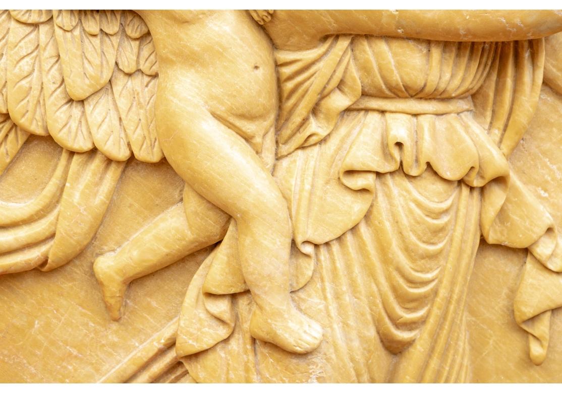 A finely carved yellow stone neoclassical roundel in relief with a flying winged female figure in flowing pleated robes with a putto holding a flaming torch and catching a ride on her shoulder. She looks back at him as she tosses roses in the sky.