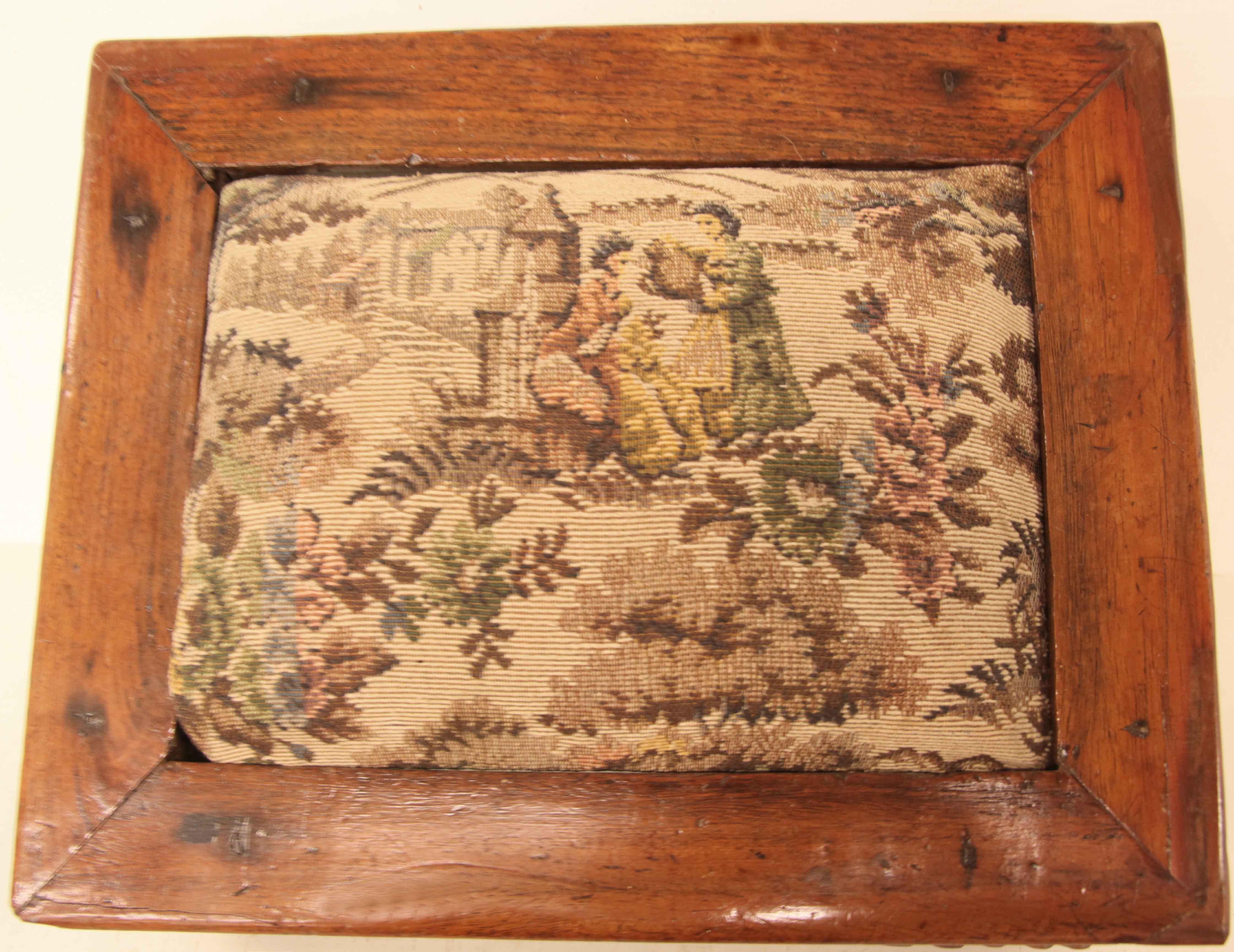 Carved teak foot stool,  the raised fabric cushion features two women at a well with flowers and foliage around them., this is followed by a 2'' wide flat surface.  The apron  has carved scrolling foliage with a central rosette on two sides.  Below