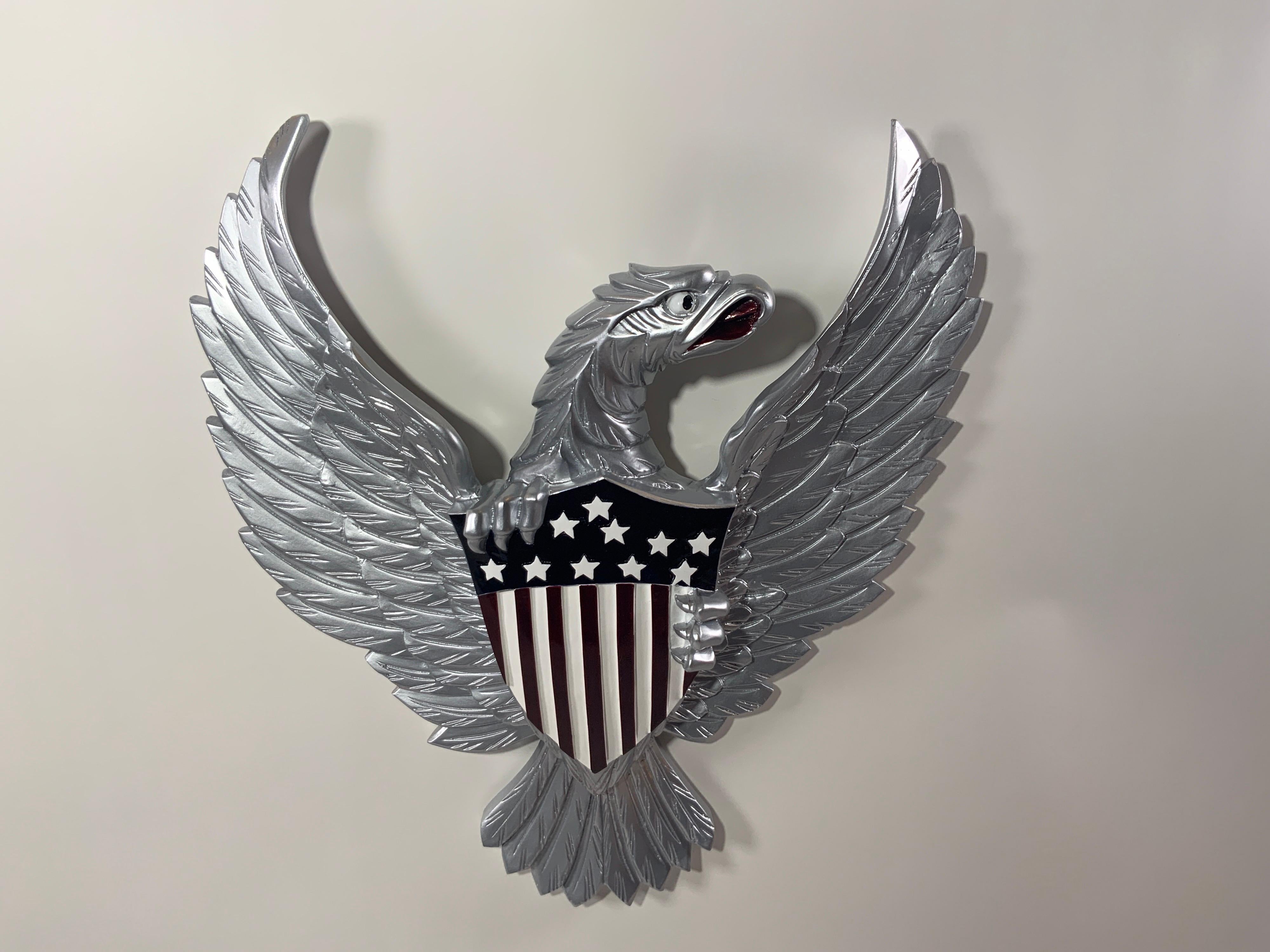 Geschnitzter Holzadler mit glänzend silberner Lackierung. Der Adler ist sehr detailliert und umklammert das Große Siegel in Anlehnung an das US-Wappen. Hinteres Aufhängeseil.

Gesamtabmessungen: 21