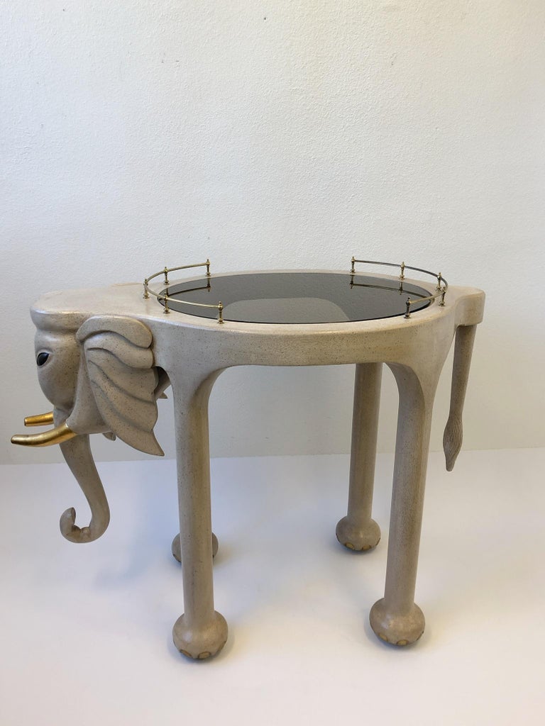 A spectacular 1990s carved wood elephant bar cart on casters by Marge Carson. The elephant is lacquered off white and gold leaf. The glass is smoked. The bar retains the Marge Carson label.
Dimension: 39.75” high, 50” wide, 26” deep.