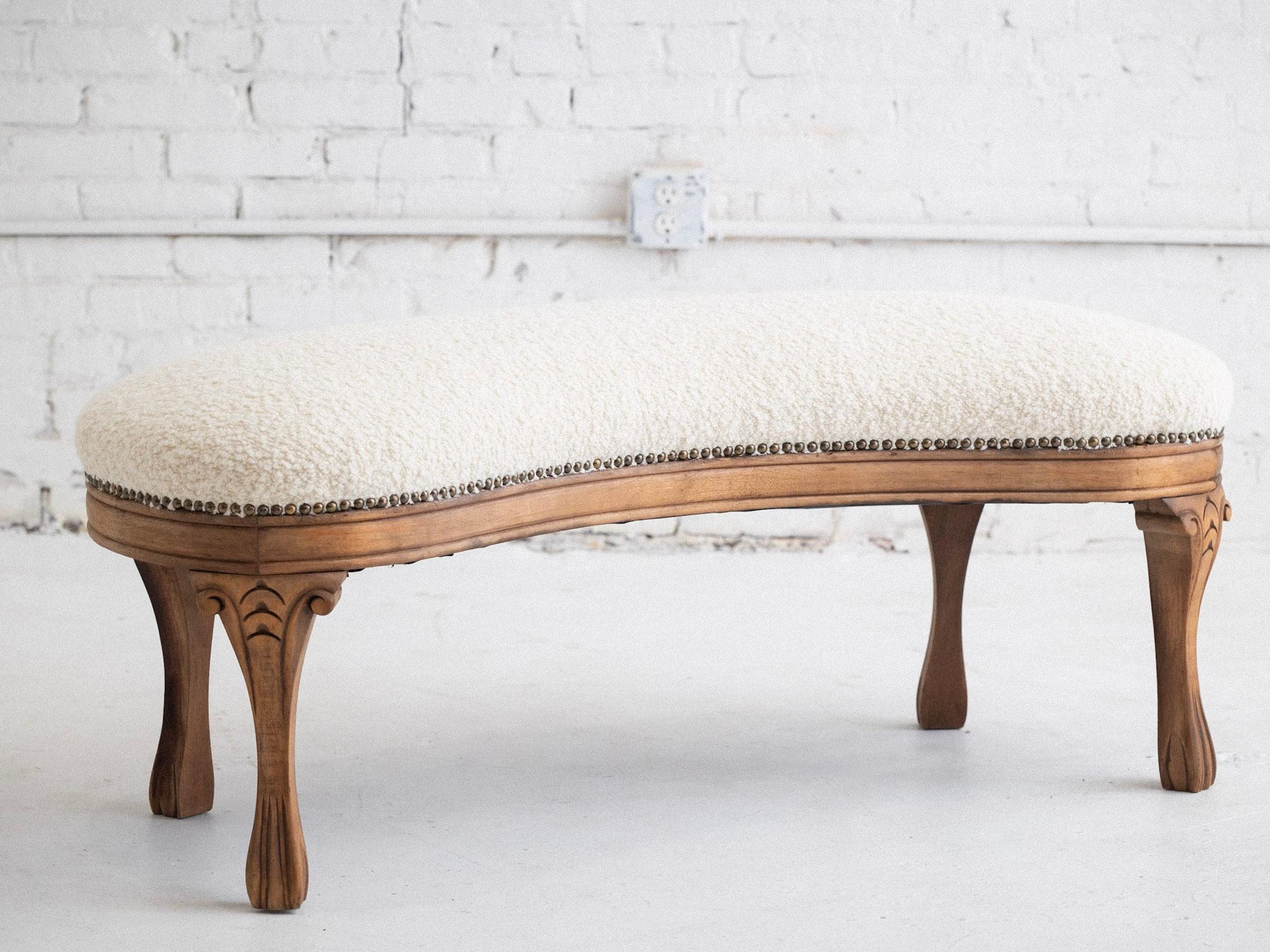 An early mid century carved wood bench. Kidney shaped silhouette with carved wood base and legs in a natural raw finish. New cream wool bouclé upholstery with bronze nail trim.