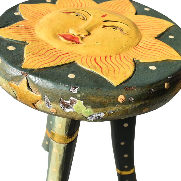 A fun whimsical carved wood side table or stool. This piece features a carved figure of the sun on its top. The sun is given life with a carved nose, lips, and eyes. She wears a red lip and has a red bindi on her forehead. The table has three legs,