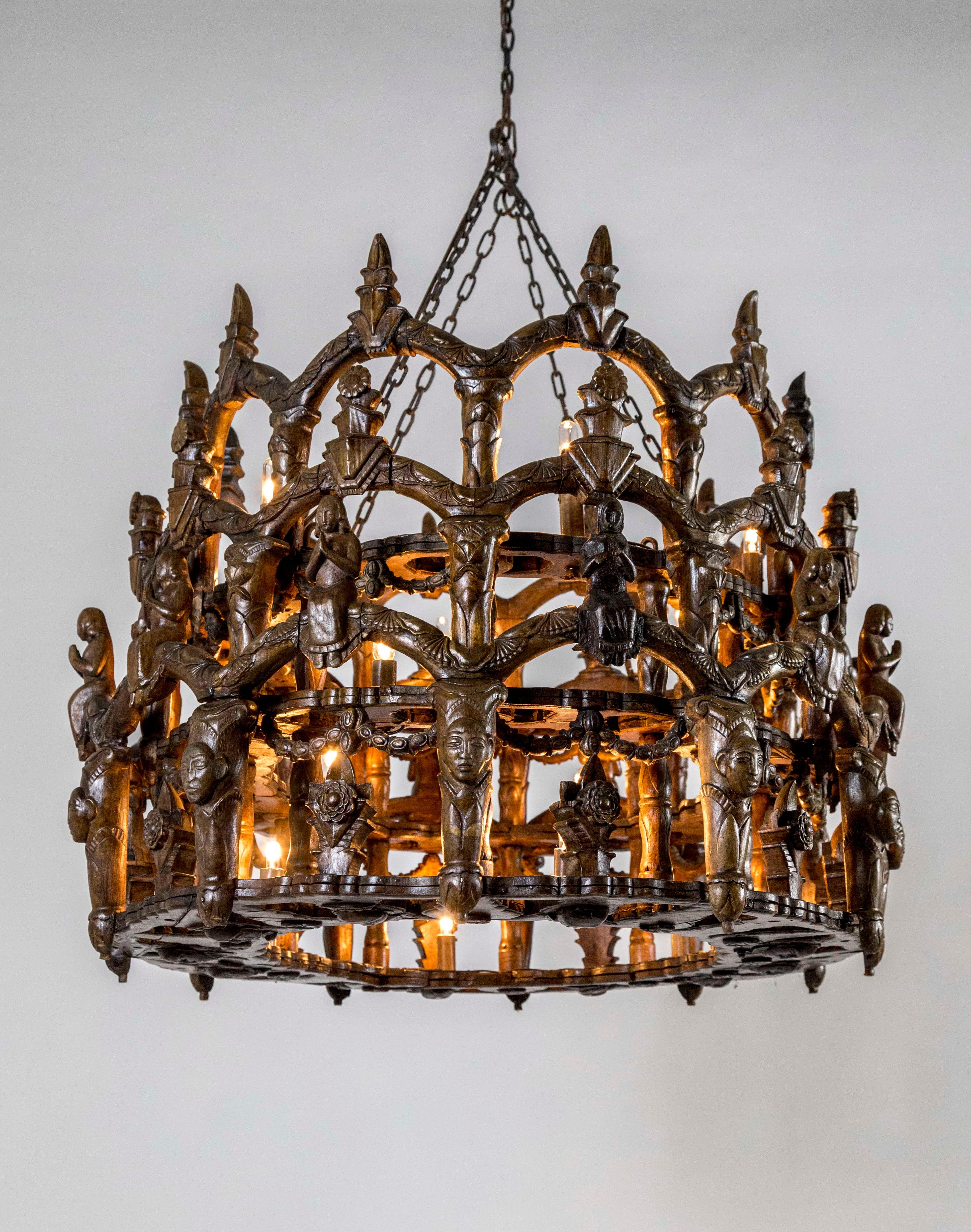 A wooden, oval shaped, folk chandelier from the late 19th century made in Central or South America with Spanish Colonial Gothic influence. The hand carved design is three tiers of arches with lotuses, flowers, praying ladies, faces, and garland. 18