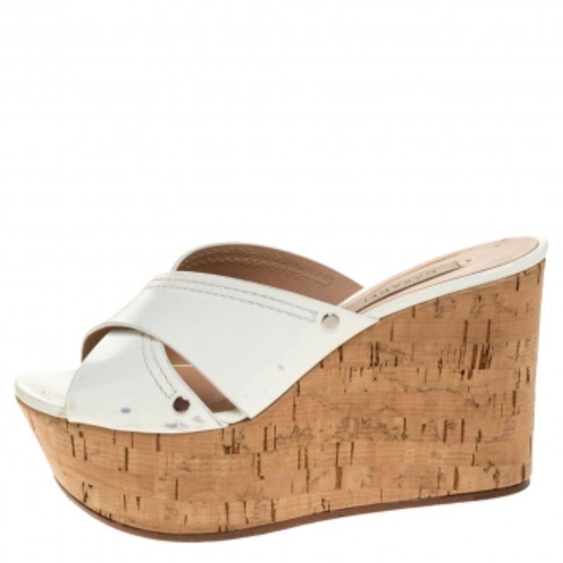 Estas elegantes cuñas proceden de la casa Casadei. Están confeccionados en charol de color blanco. Tienen puntera abierta, tiras cruzadas y tacón de cuña de corcho de 9,5 cm. Están acabados con suelas duraderas.