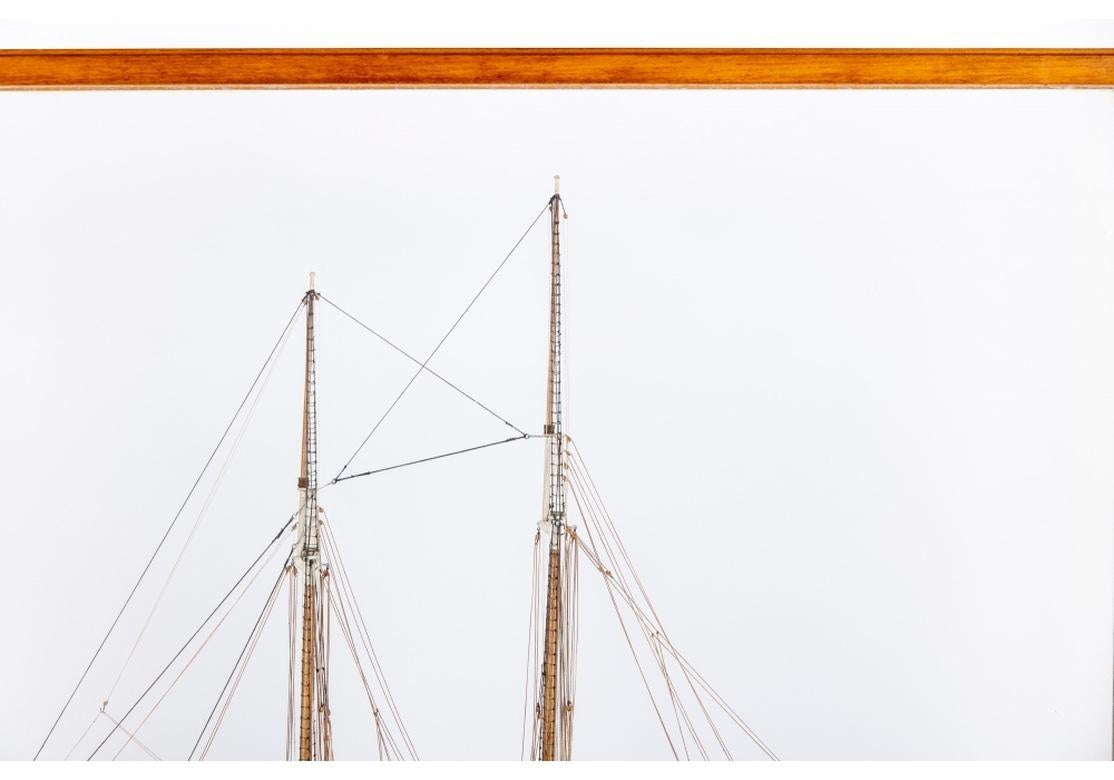 Finely crafted and minutely detailed two masted schooner in wood with a black painted hull and with string rigging, anchors, hatches, boats, barrels and some fine chains. The Scale is 1/8th inch = 1 Foot. Mounted on a base within a glass and wood