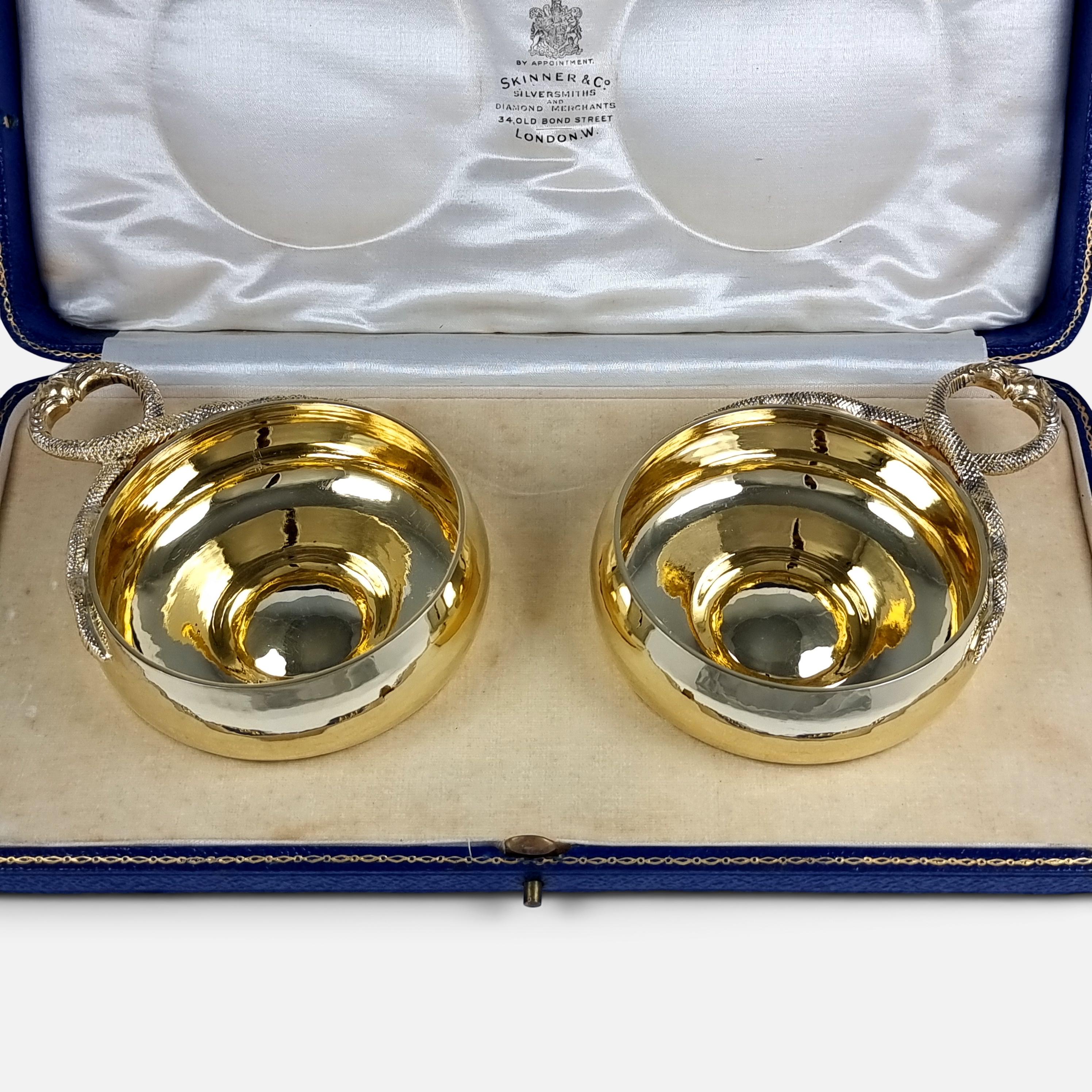 A cased pair of George V sterling silver-gilt wine taster cups. The wine taster (otherwise known as tasse de vin) cups are of round form, with spot-hammered finish, and each with a single snakehead handle. 

The wine tasters are stored in their