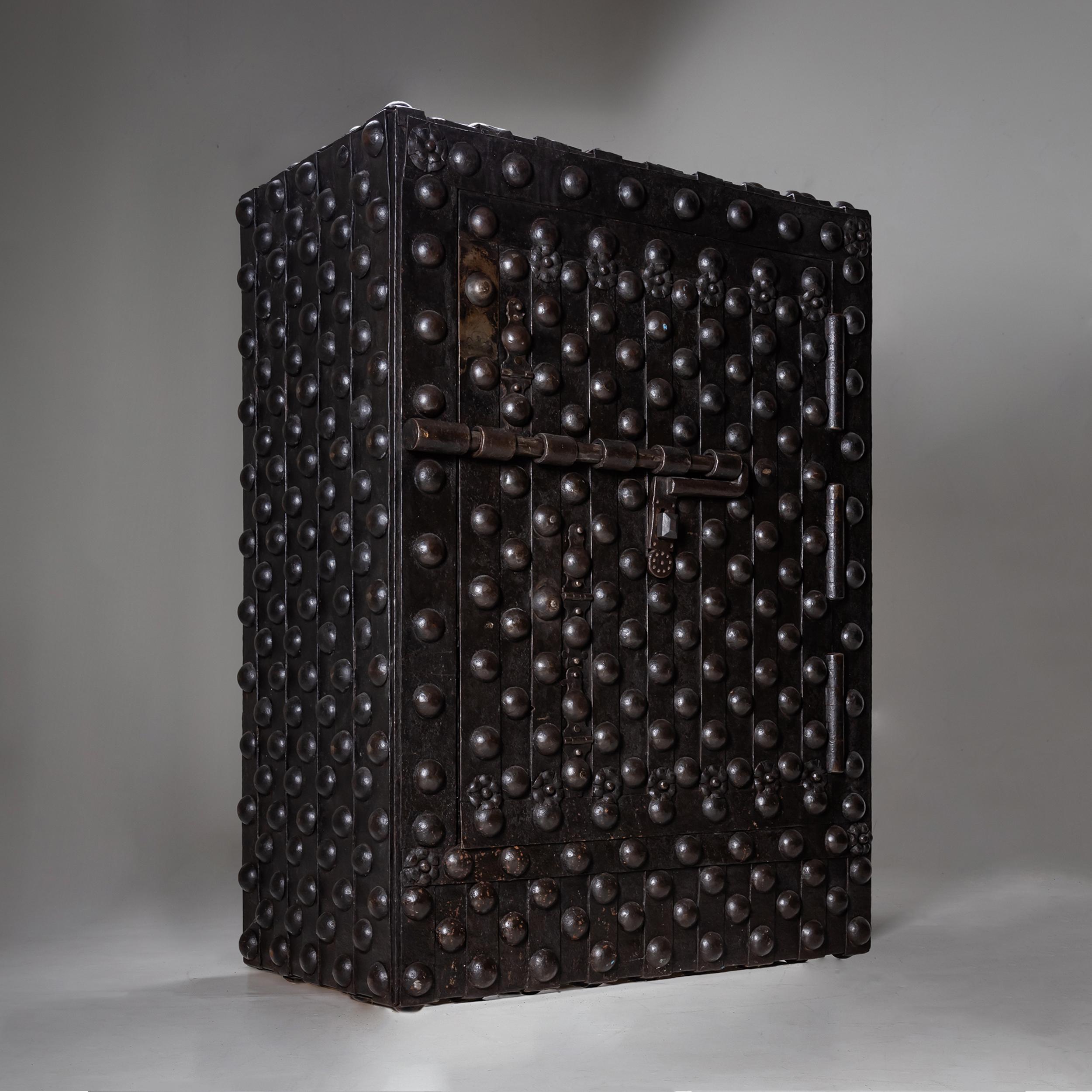 Large Italian black cassaforte from the 17th century, featuring heavy iron bands and rivet detailing. The door is adorned with decorative rosettes, while large strap hinges and a robust latch define the striking front.
The safe is opened via an