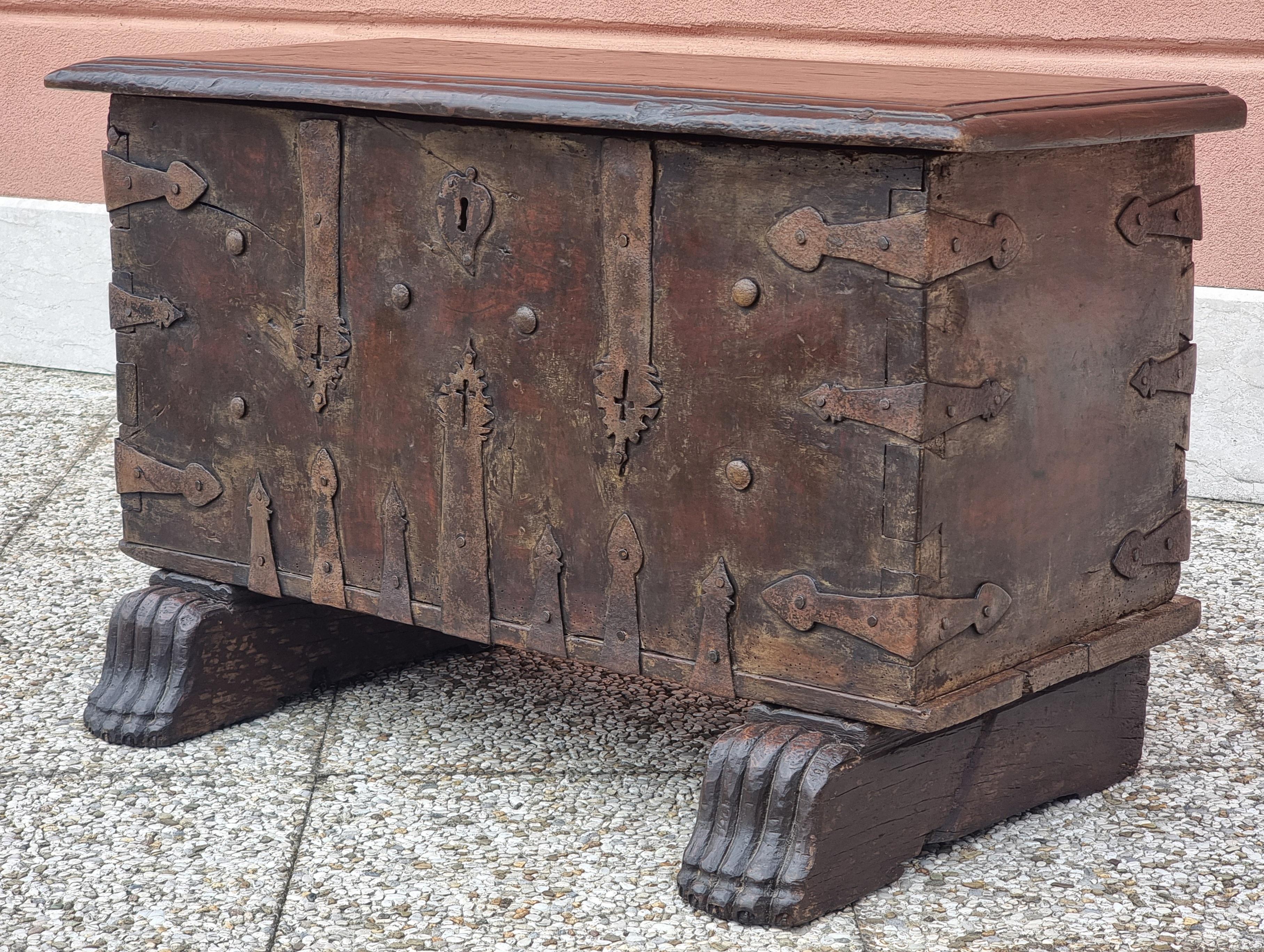 Pequeño arcón-banco de madera maciza de nogal, reforzado y decorado con llamativos herrajes de hierro forjado, consistentes en largas placas en forma de lanza y espárragos forjados, muchos de ellos perforados con cruces o grabados con motivos