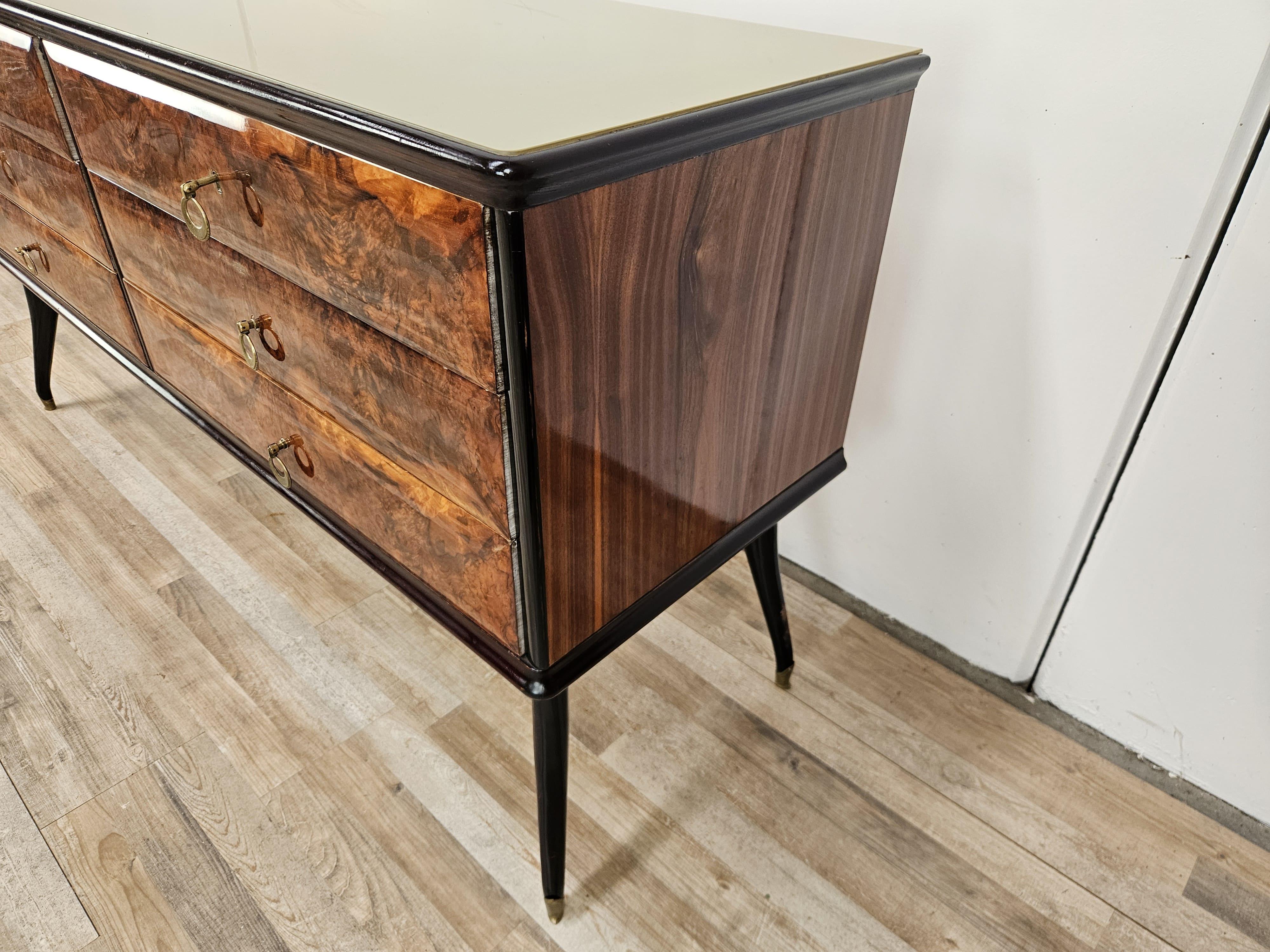 Elegante cómoda italiana de mediados del siglo XX revestida en burl de caoba con elegantes tiradores de latón y tapa de cristal original de época.

Destaca la línea de las patas con las escuadras de latón, que recuerdan un diseño moderno y