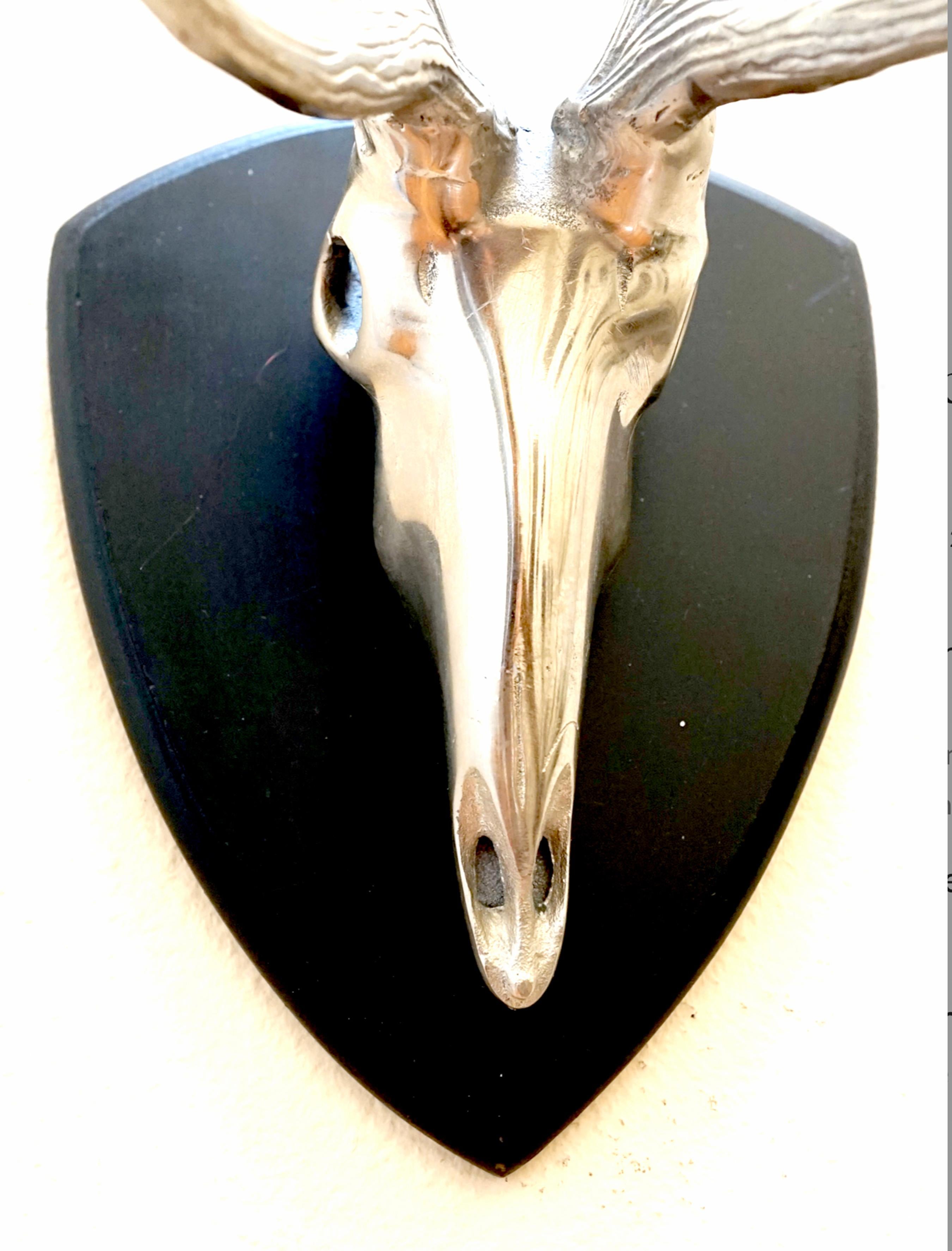 The sculpture is a vintage cast metal antelope skull wall ornament. It is unattributed, and was made in the second half of the 20th century. 
The piece is sleek, mounted with two three-point antlers on a shaped three-sided wooden base, unmarked. It