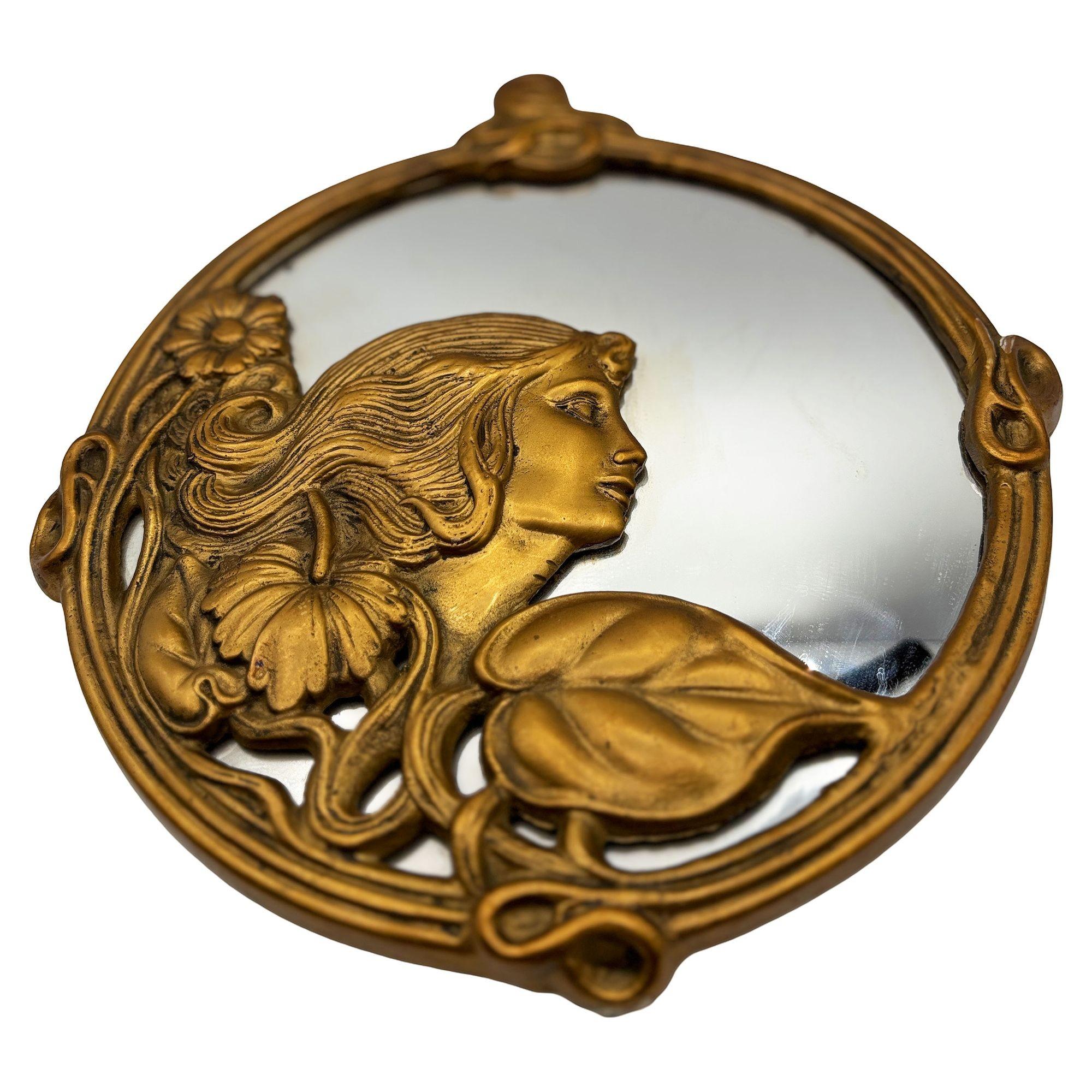 Specchio da parete rotondo in ottone fuso e metallo dorato in stile Art Nouveau con un ritratto femminile circondato da un motivo floreale di edera che circonda lo specchio. Lo specchio ha una forma stravagante e fluida che caratterizza il deco di
