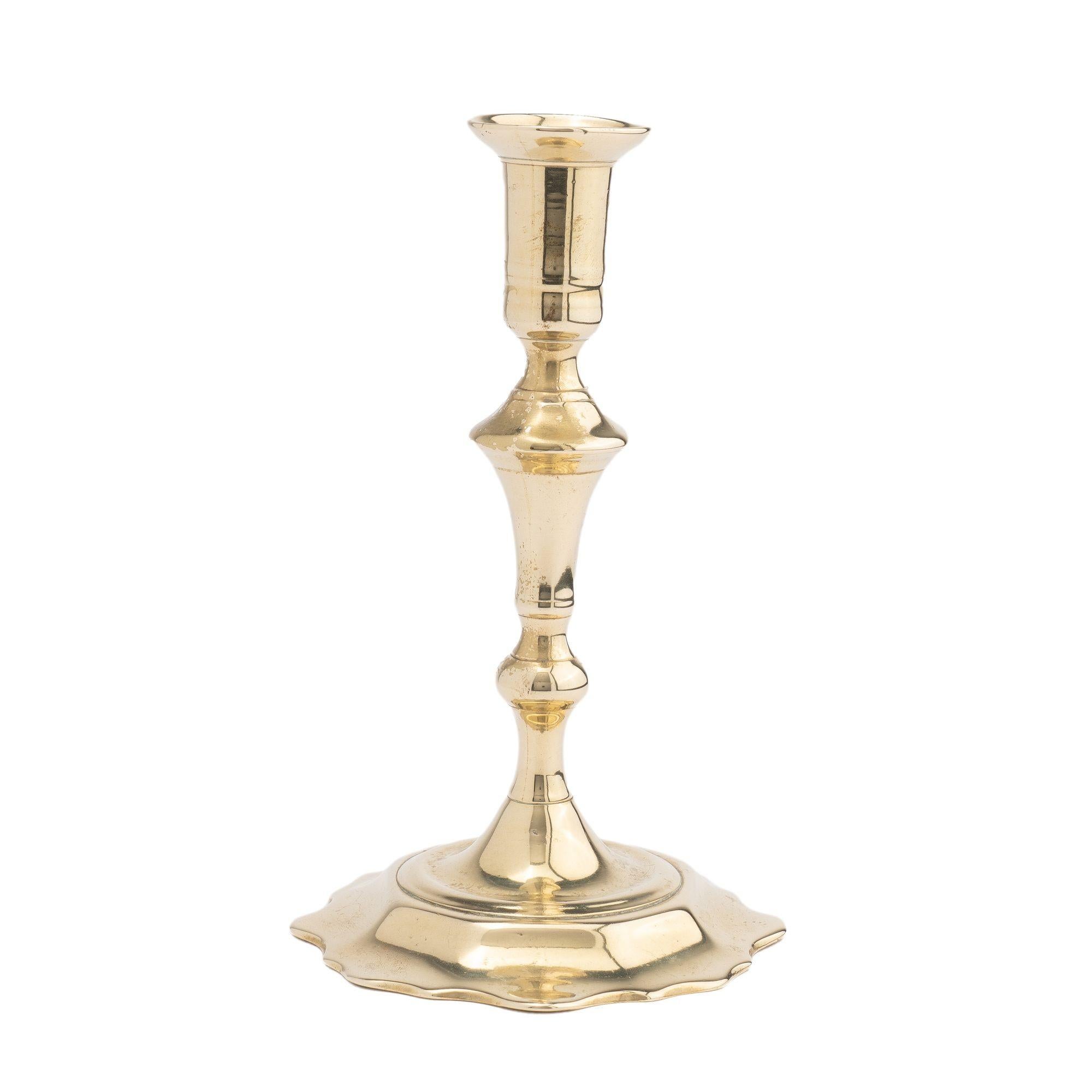 Queen Anne scollop base candlestick. Seem cast two part cast brass candlestick shaft. The shaft supports a cylindrical candle cup with dished circular bobeshe. Below the cup is a suppressed knob on an inverted trumpet form, smaller knob and short