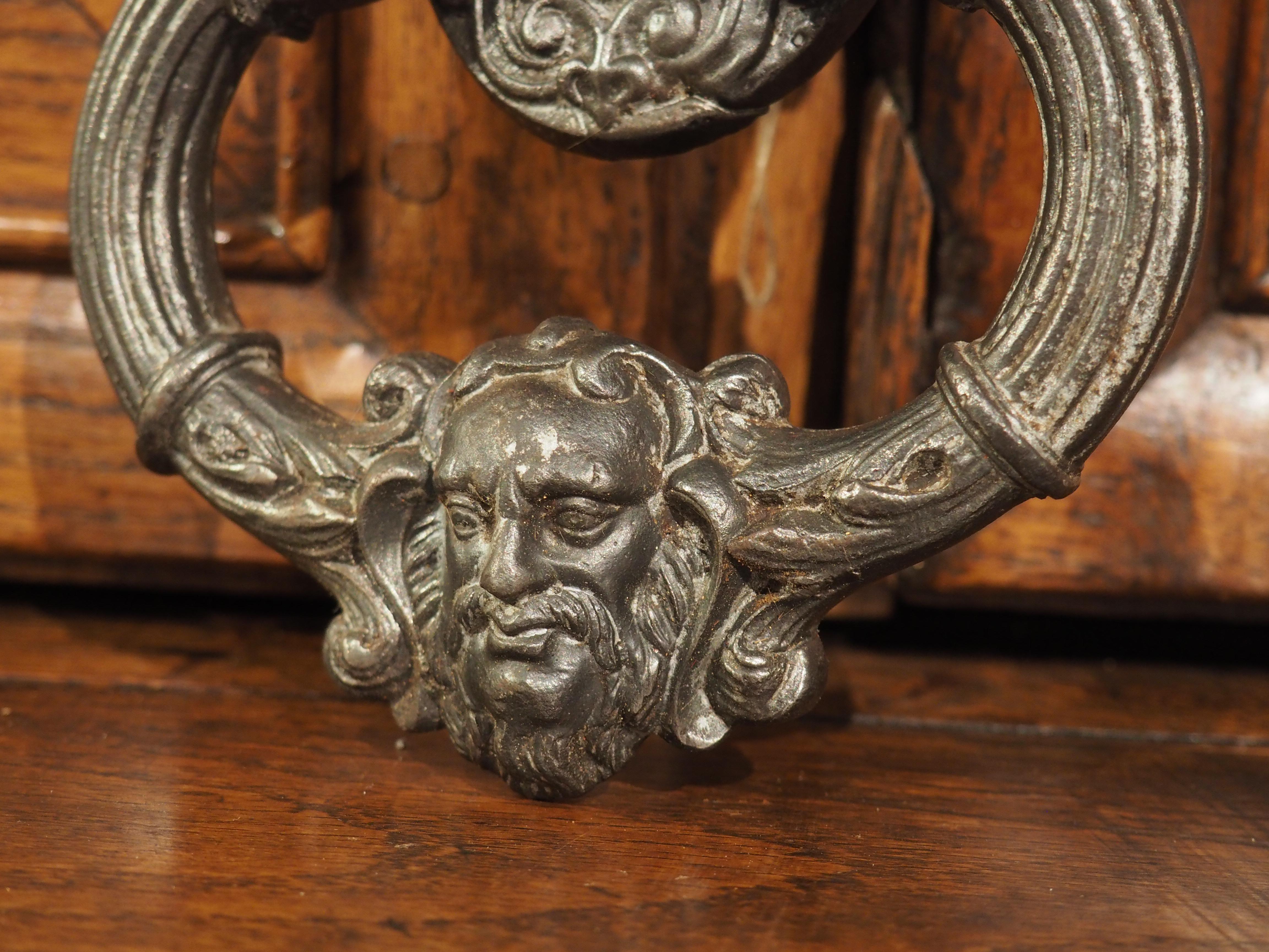 Cast in iron during the 1900s, this mascaron knocker will instantly enhance any front door. Its oval backplate is exquisitely adorned with intricate scrolls, delicate foliage, and a central rosette. The knocker’s shapely drop handle, enriched with