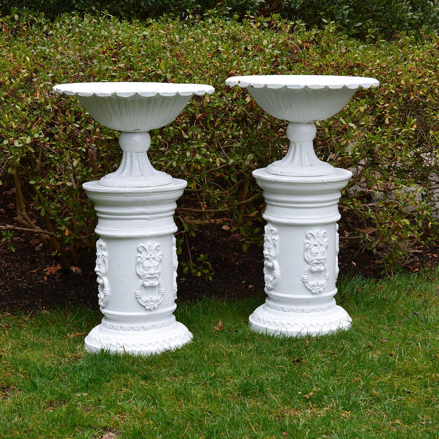 A fine pair of cast iron tazza form urns with fluted foot and scalloped rim, English, circa 1930, on associated pair of cast iron pedestal columns with masks. Measures: Overall height 36.5 ins. The urns 13 ins. high, 22 ins. overall diameter, the