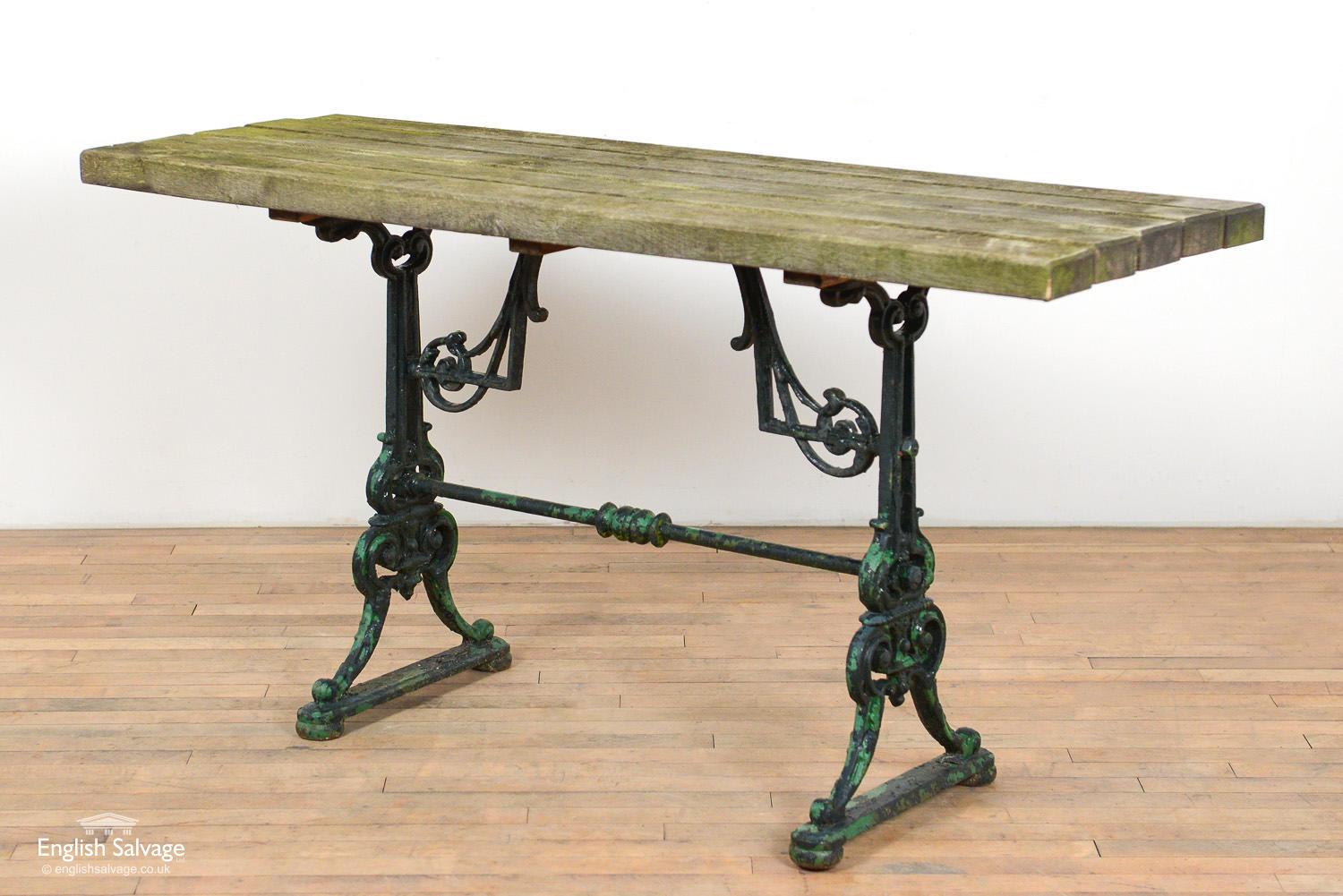 Salvaged table with cast iron base in ornate scrolling and angular design. Peeling paint remnants give it a weathered patina. Rustic top made up of five narrow and thick boards.