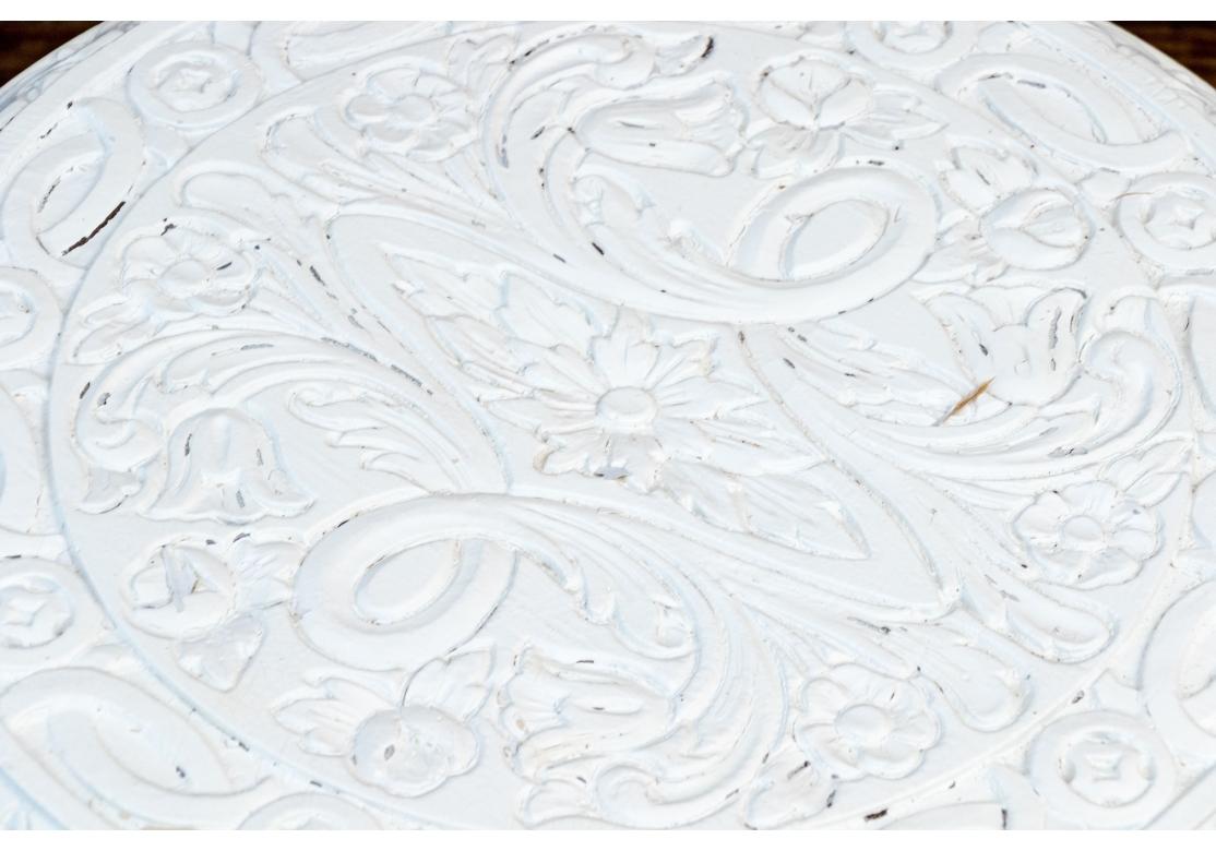 La petite table ronde d'accent oriental  peinte en blanc et que l'on pense être en résine synthétique coulée. Le plateau est orné de rinceaux sculptés sur une frise avec un bandeau décoratif sculpté. Elle repose sur quatre pattes en forme de tête et