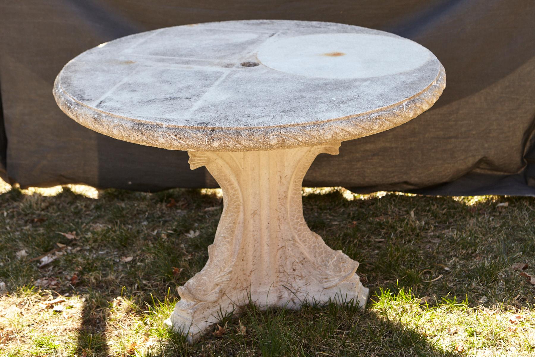 Round cast stone center table bench with an ornamental carved band of alternating concave and convex sections around the edge, reminiscent of waves, mounted on a triangular base with decorative fish/koi motives.
The top sits firmly on a hollow iron