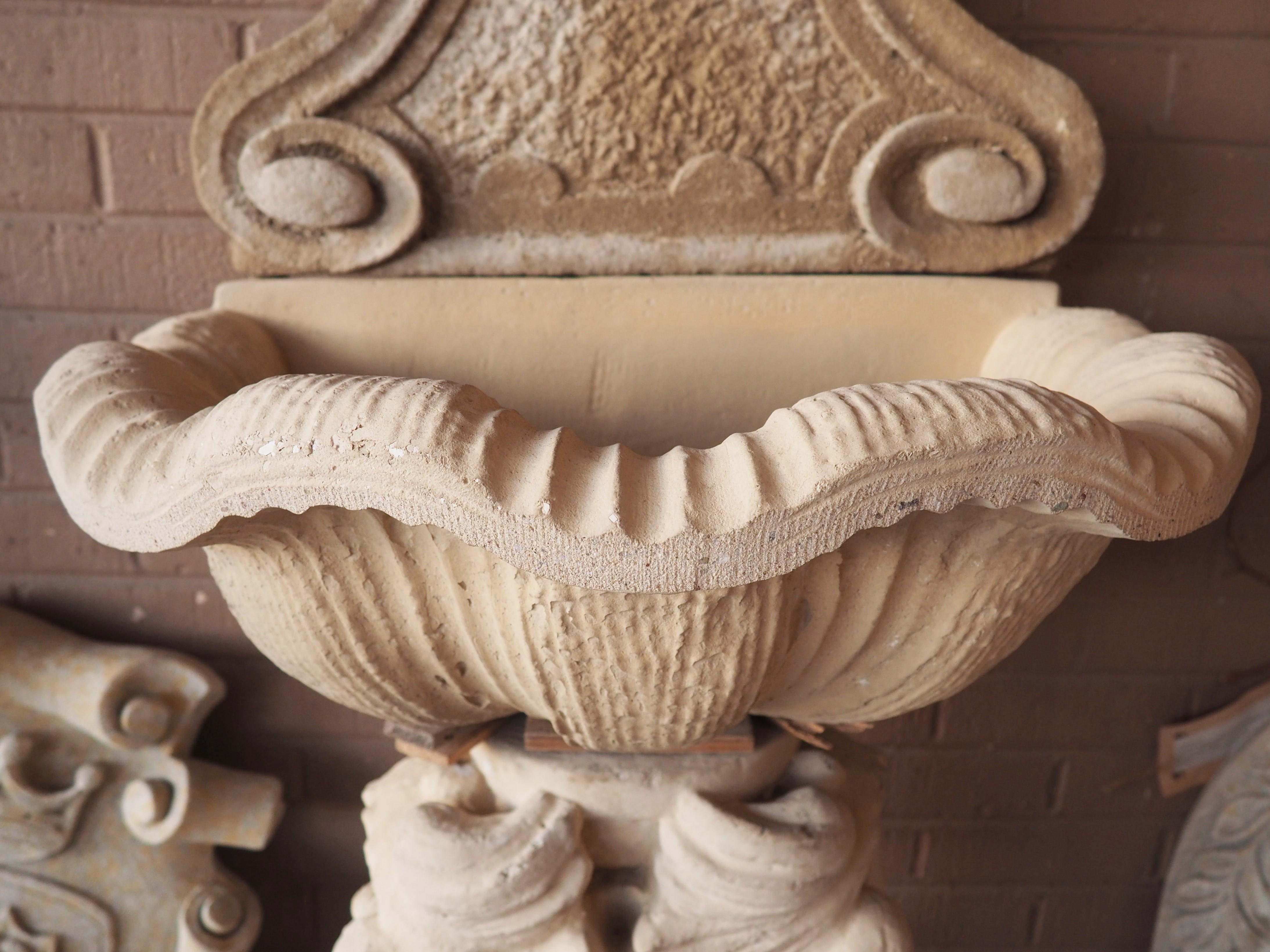 A charming French wall fountain comprised of three pieces of reconstituted, or cast stone, has decorative motifs of a putto and a pair of dolphins. A pedestal formed by two intertwined dolphins supports a scalloped shell basin, which is topped by a