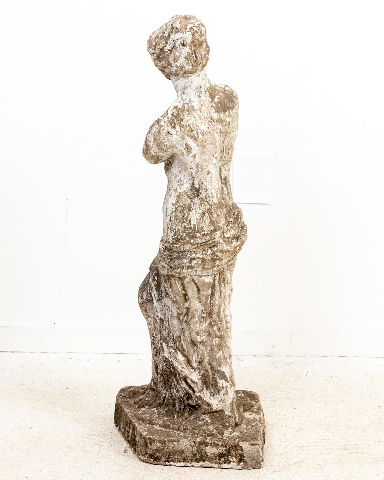Classical style cast stone garden statue in a weathered finish depicting a robed, demi nude female figure on plinth in the manner of the Venus De Milo statue. Please note of wear consistent with age and exposure to the elements such as