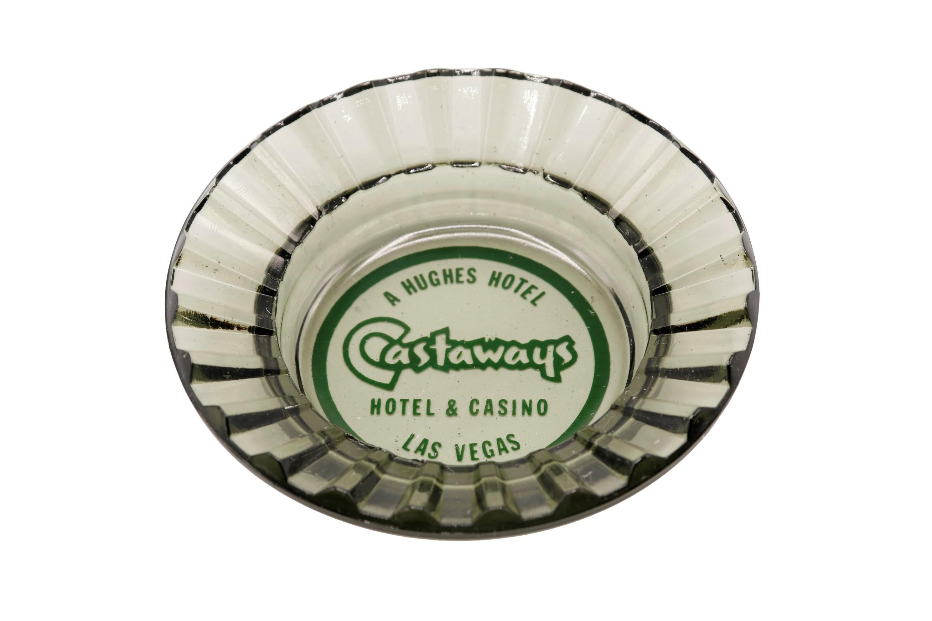 A gray tinted glass ashtray from The Castaways Hotel, a hotel and casino in Paradise, Las Vegas, Nevada. The center is printed with a green circle branded with the Castaways logo. The hotel was opened in 1963, purchased in 1967 by billionaire Howard