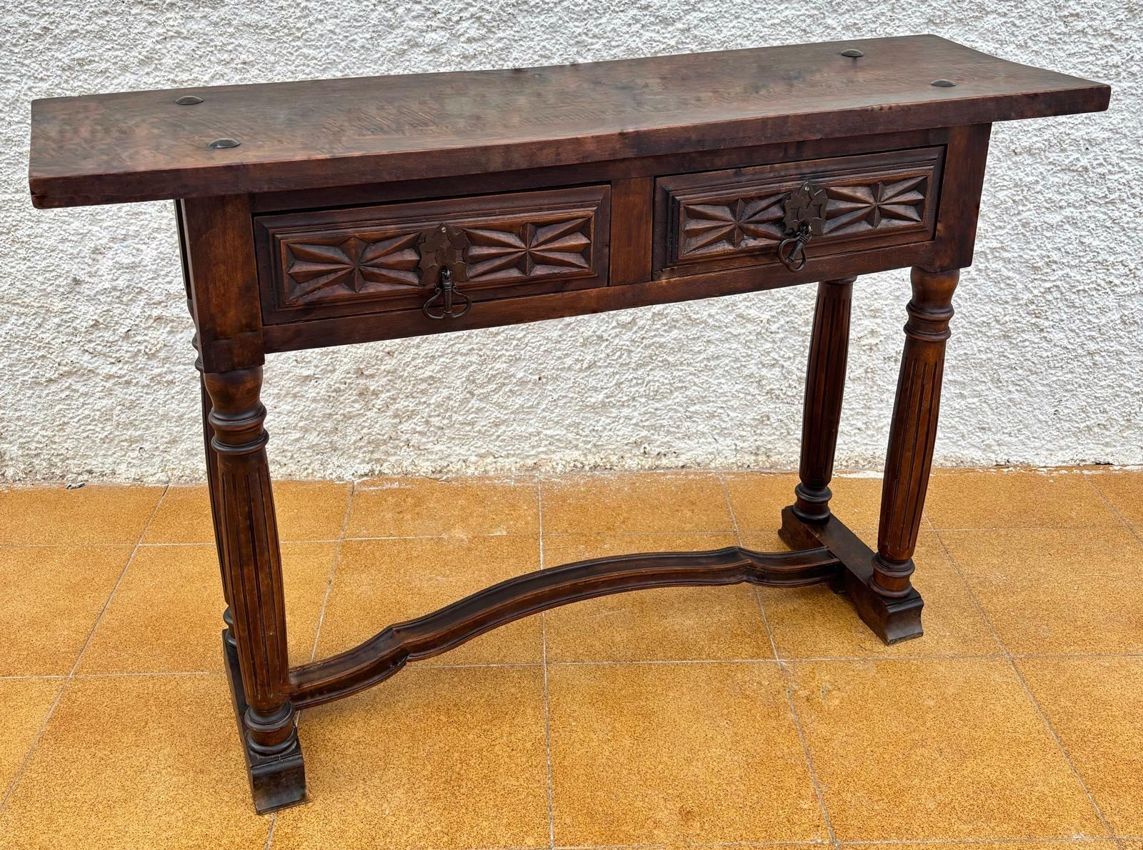 Consola castellana con dos cajones tallados, con tiradores de hierro que se apoyan sobre cuatro patas en forma de columnas abovedadas y a su vez talladas con líneas rectas. Estas cuatro patas están unidas por un cierre arqueado de madera.
