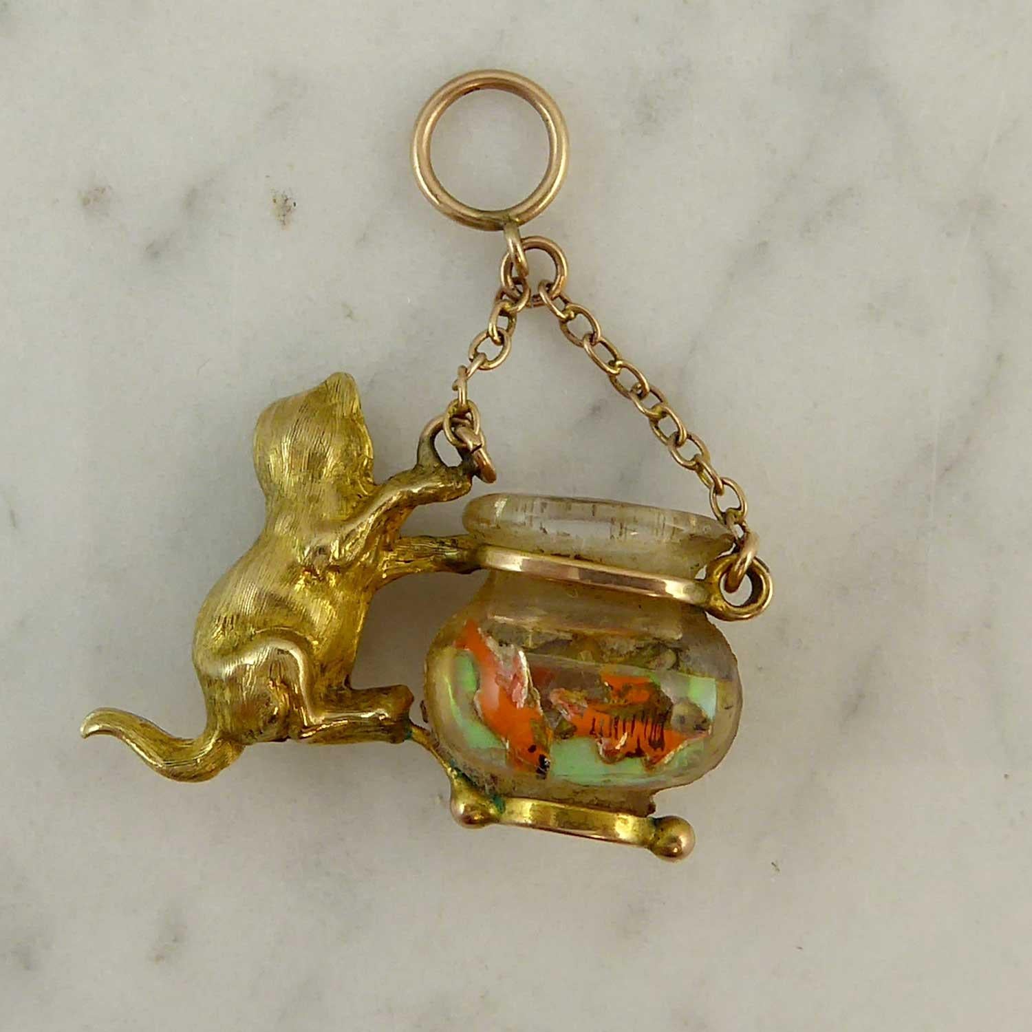 An unsual charm dating from the mid-20th century showing a naughty, green-eyed cat climbing up the side of a large bowl in which there are two unsuspecting goldfish. The charm suspends from two strands of gold trace chain leading to a pendant loop.