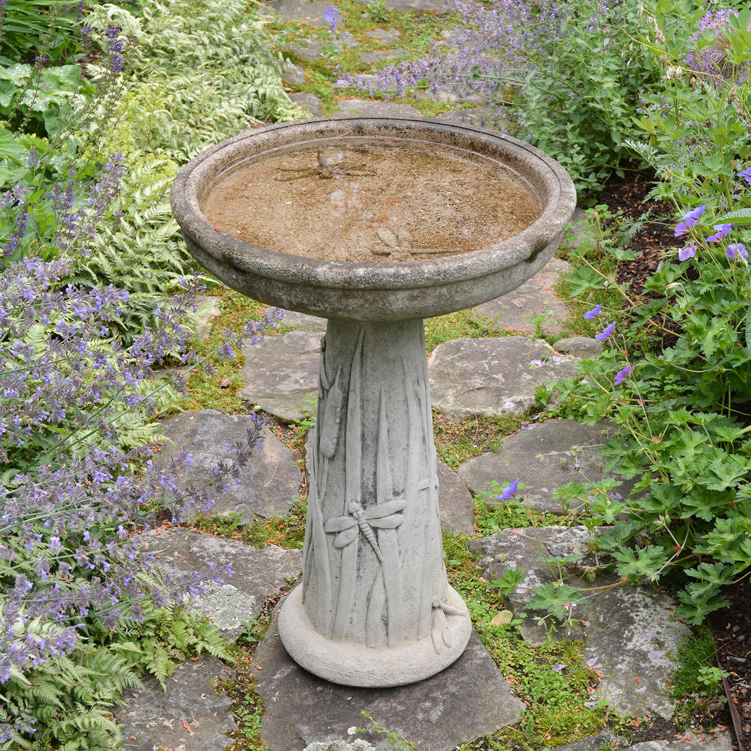 A composition stone birdbath with cattail and dragonfly motif on pedestal and in bowl.