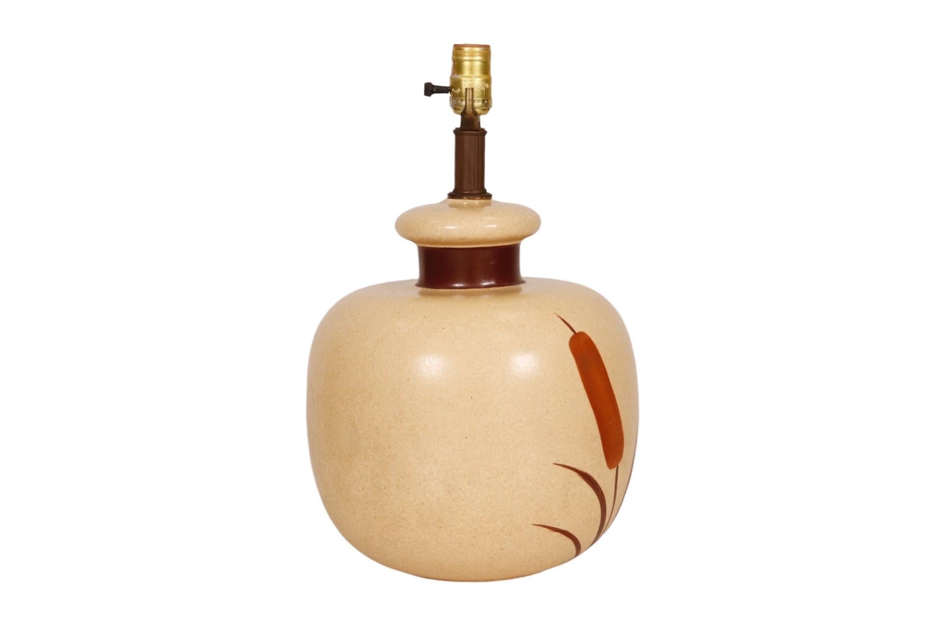 A ceramic gourd shaped table lamp in a lightly speckled oatmeal glaze, decorated with hand painted caramel colored cattails on the body. The neck of the lamp is painted a burnt umber brown to match the leaves.
  
