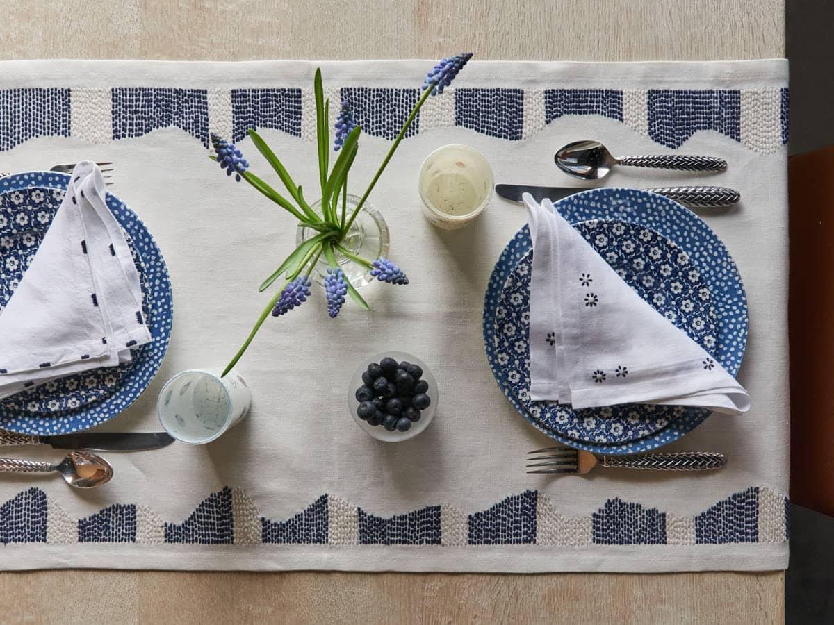 A fairy-tale table. White linen table runner with artisanal hand embroidered navy and cream flowers to dress the table and turn every occasion into a special one. 100% linen. Size: 50 x 160 cm.