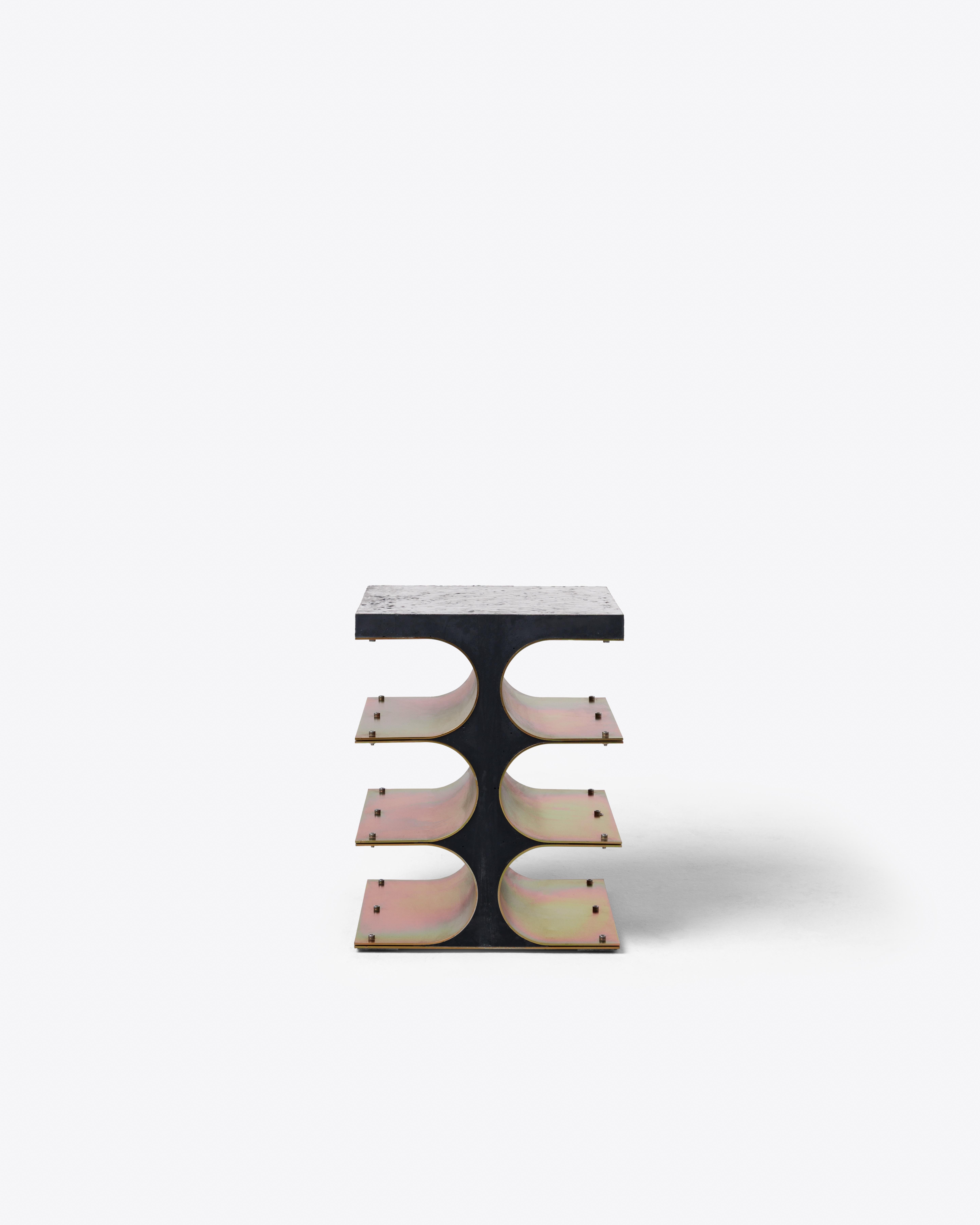 This side table combines a dense black frame with three curved shelves in iridescent steel, forming a grid of color and shadow. The contrast between opacity and shimmer reflects Simões’ architectural approach, where exposed
structure becomes central
