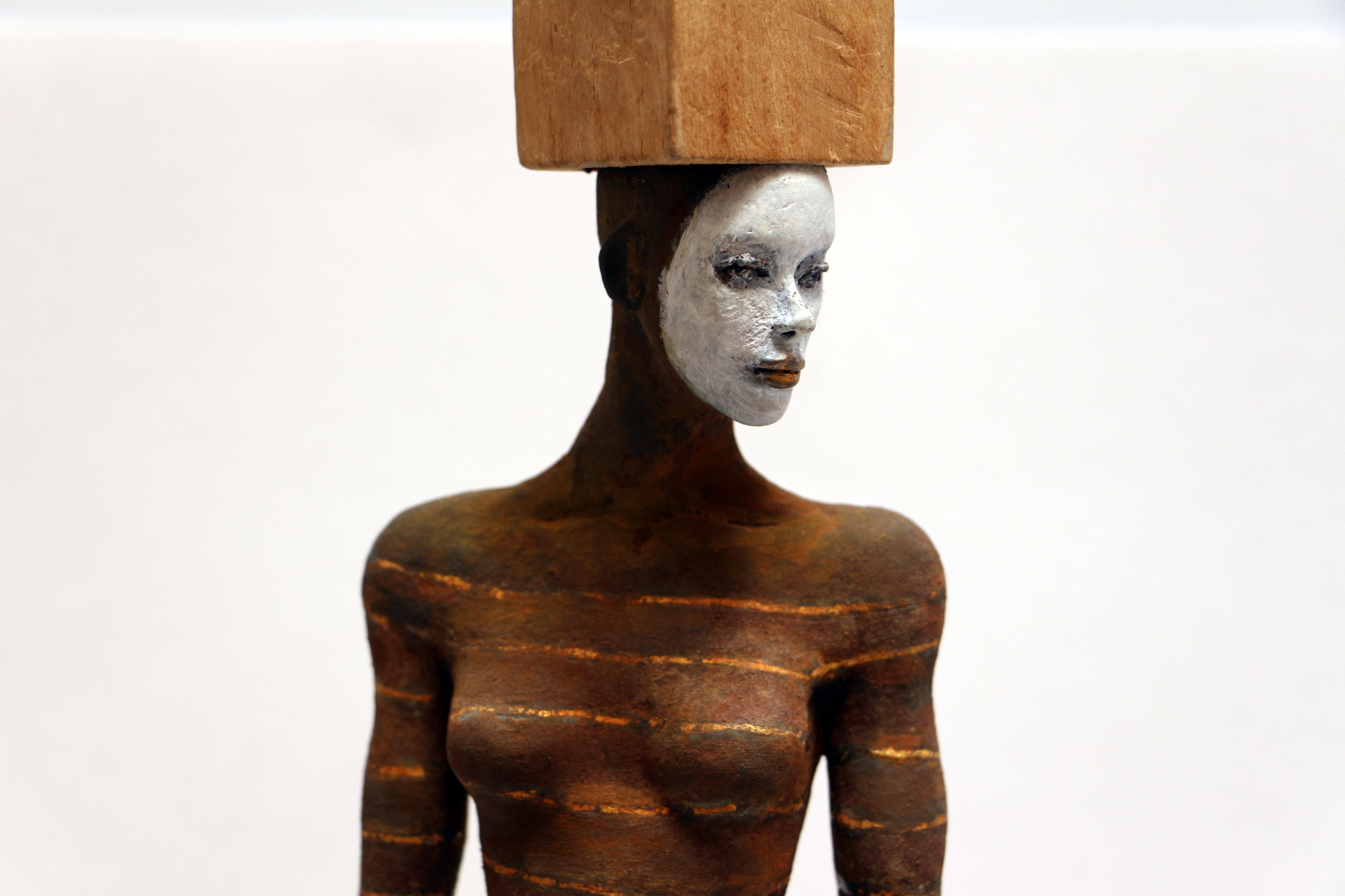 This wood and bronze sculpture depicts a nude female figure with a painted white face and horizontal stripes along the body. She is standing on a base, and wears a head accessory that consists of an open structure with small spherical shapes.