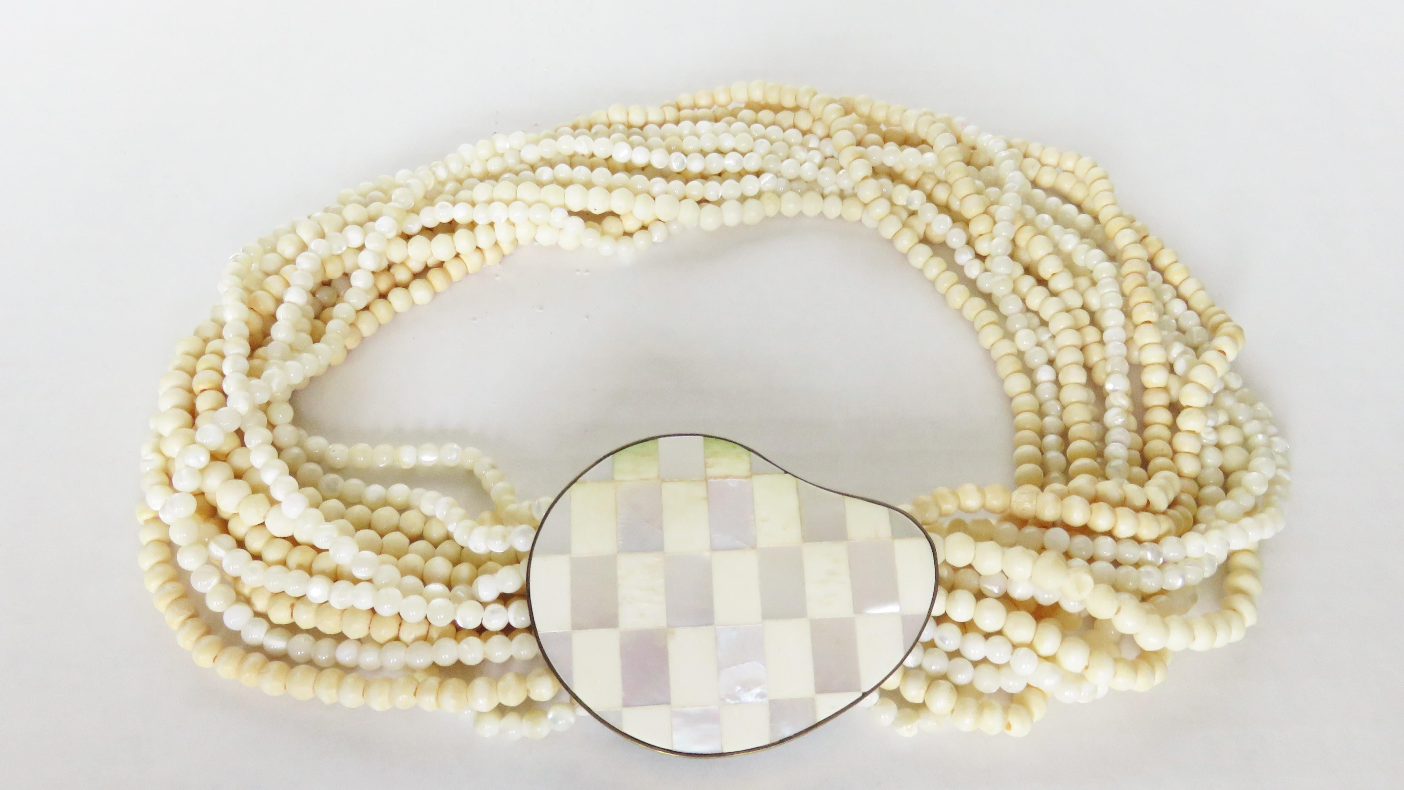 Magnifique collier ras de cou torsadé de l'artiste Celia Sebiri composé de 12 brins de perles de nacre et d'os reliés par un grand fermoir incrusté de nacre de 2,25 X 1,34 pouces. Il se ferme à l'aide d'un double crochet en laiton sur le dessous et