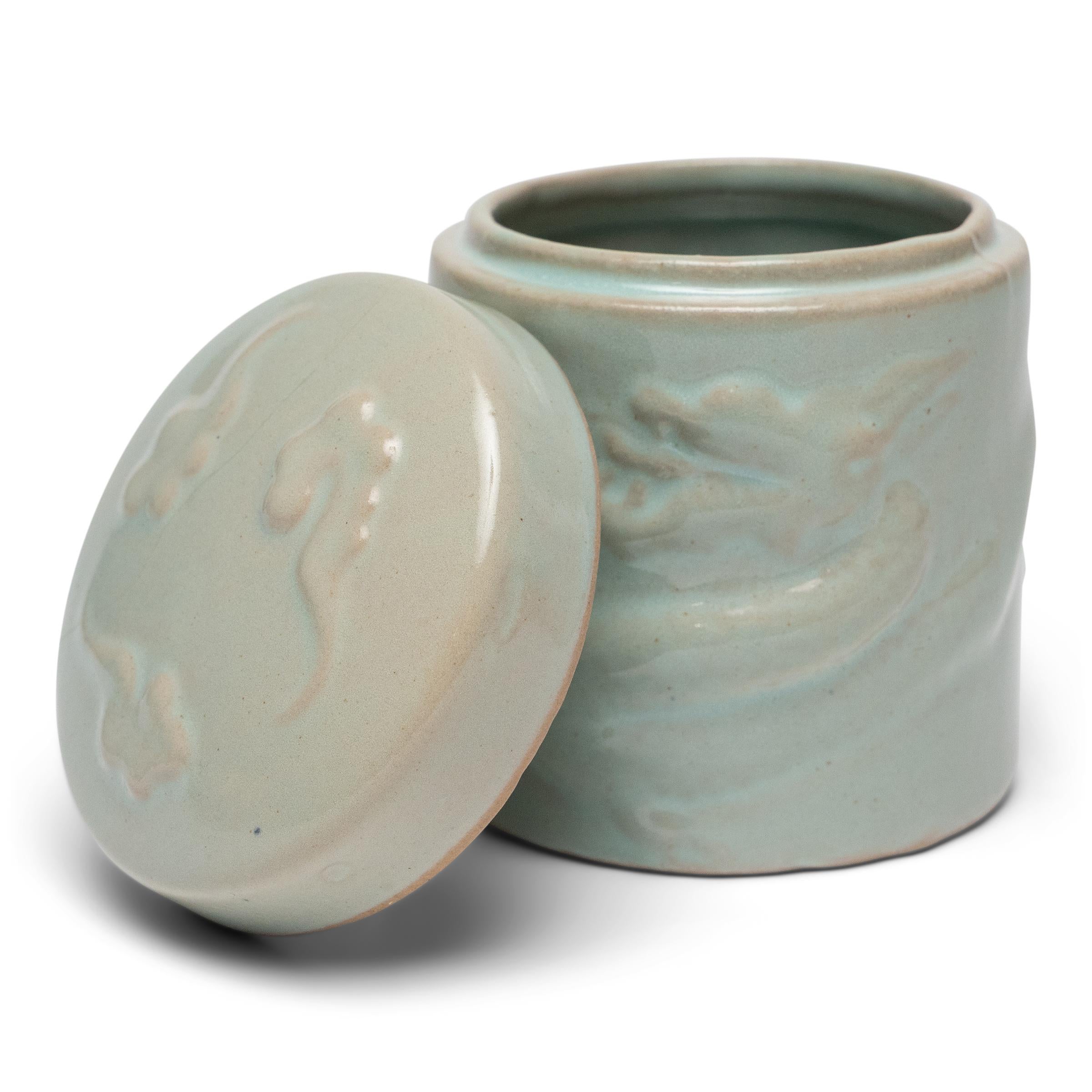 Cette petite jarre en céladon a une forme cylindrique simple, traditionnellement utilisée pour conserver le thé en feuilles ou d'autres herbes séchées. Trois petits nuages décorent le couvercle rond et un dragon céleste serpente en bas-relief sur