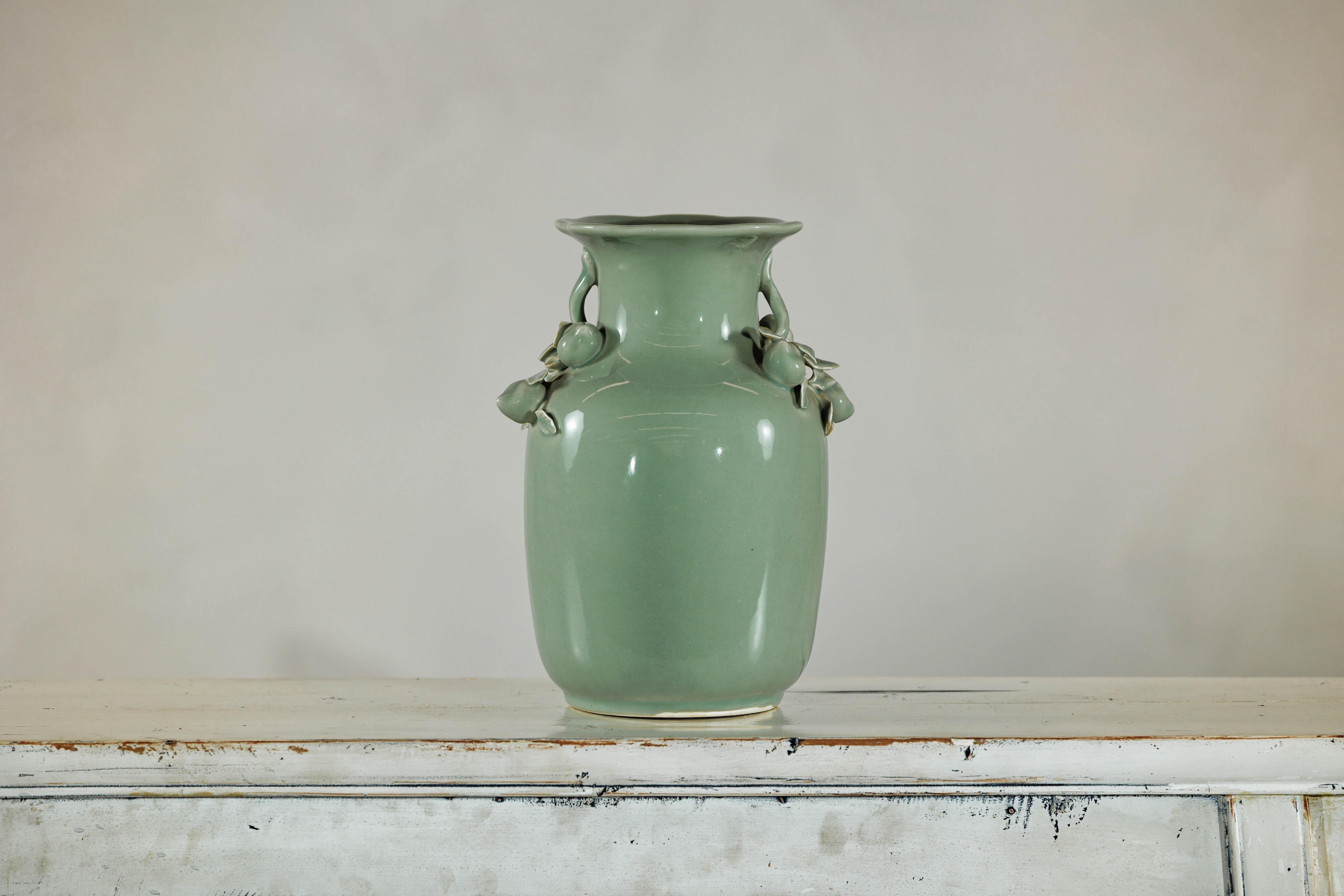 Ce vase vintage en céladon glacé présente une silhouette doucement arrondie et se distingue par un décor appliqué de fruits et de feuillages sur l'épaule. Des branches courbées émergent symétriquement du cou, chacune portant des grappes de fruits et