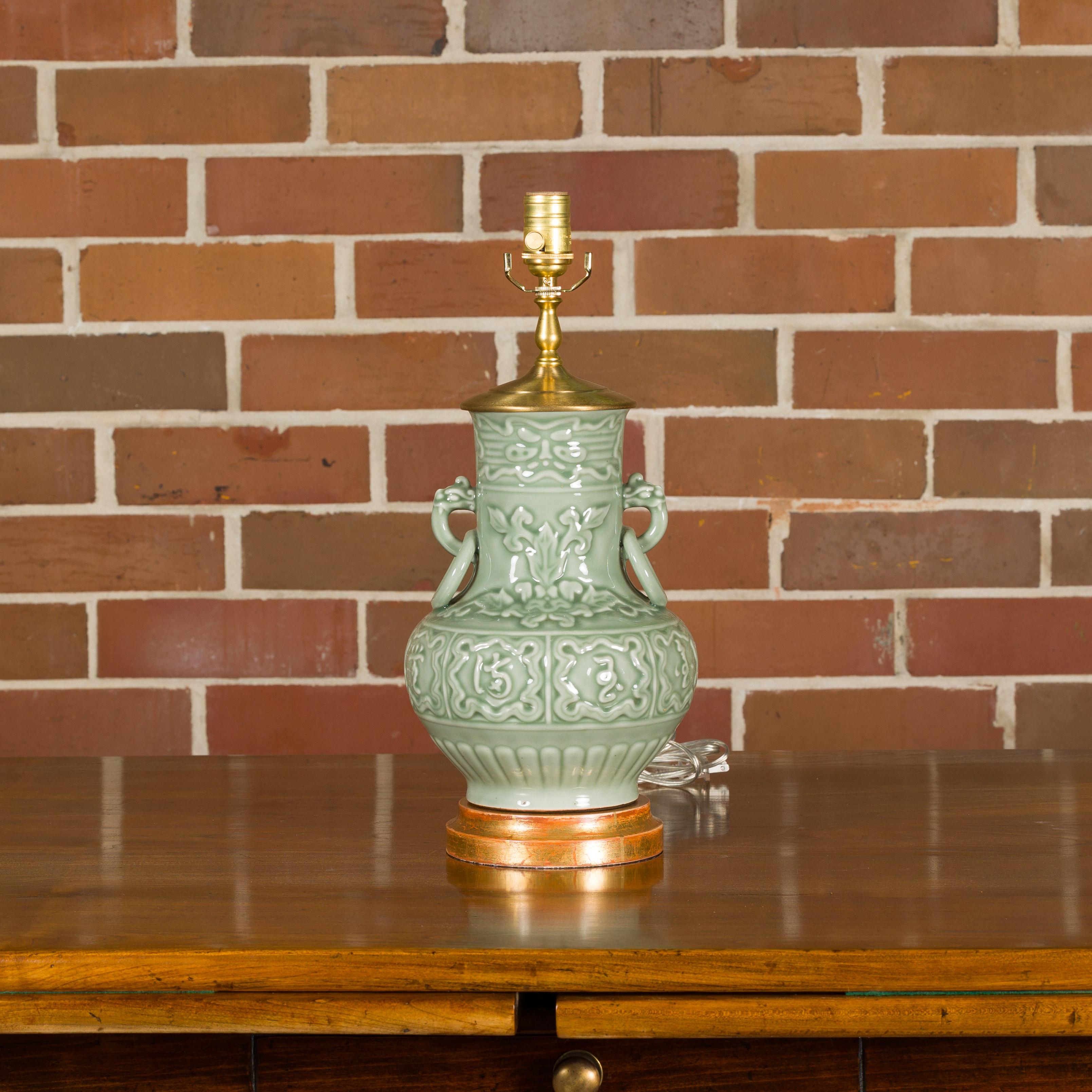 A celadon colored table lamp with raised motifs including foliage and calligraphy, ornate lateral handles and mounted on circular giltwood base. This celadon-colored table lamp is a true testament to the elegance of Asian design. With its exquisite