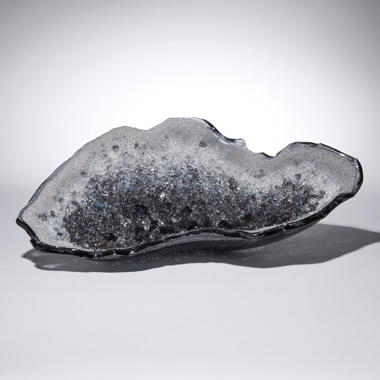 Celestine I is a unique grey, aqua & clear glass sculpture / centrepiece by the British artist Wayne Charmer. Taken inspiration from the moment that a mass of rock is literally cracked open, to reveal a carpet of sparkling gems, Charmer has filled