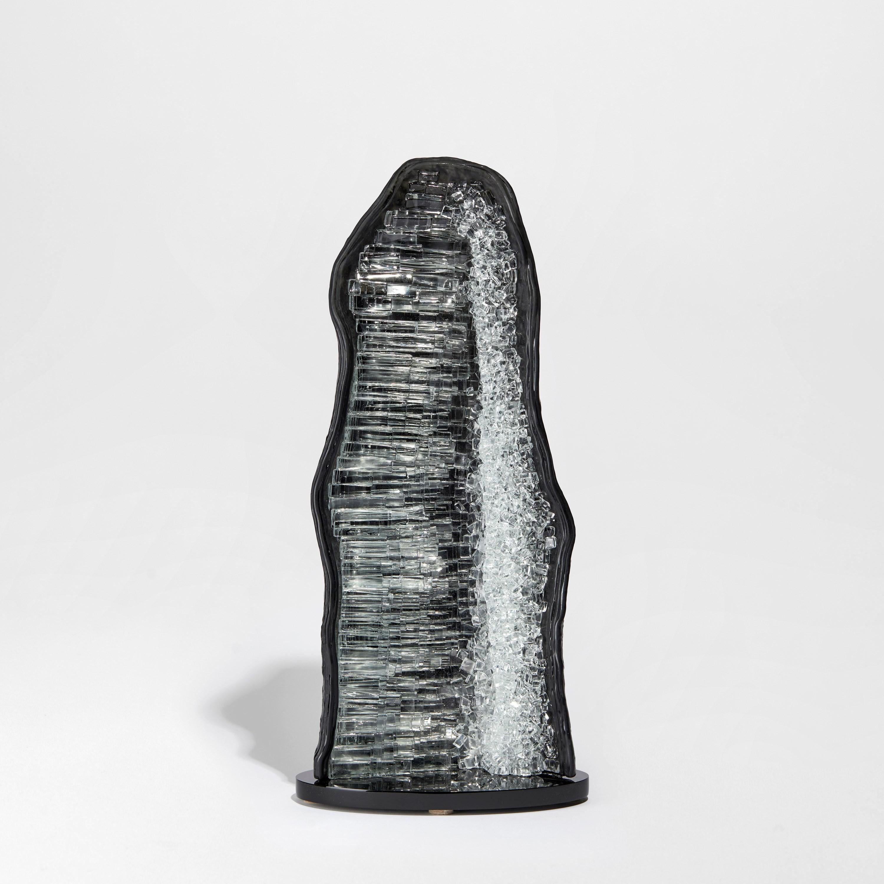 Celestite III ist eine einzigartige graue und klare Glasskulptur des britischen Künstlers Wayne Charmer. Inspiriert von dem Moment, in dem ein Gesteinsbrocken buchstäblich aufgesprengt wird, um einen Teppich aus funkelnden Edelsteinen freizulegen,