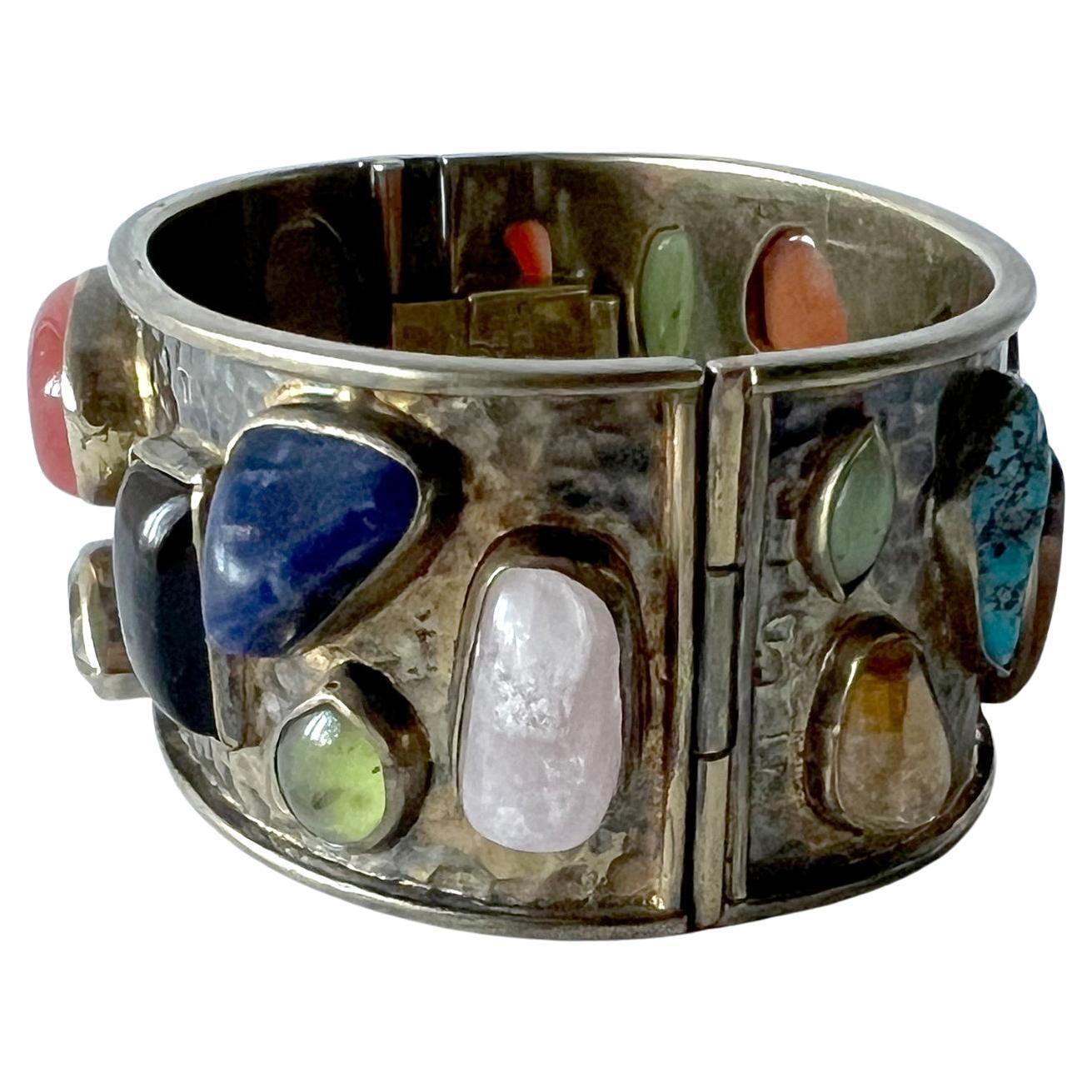 Multi colored semi-precious gemstones of turquoise, opal, amethyst, carnelian and others, set in a sterling silver hinged cuff and created by Celia Harms of Mexico.  Each stone is bezel set within a hand hammered silver, gold washed bracelet. 