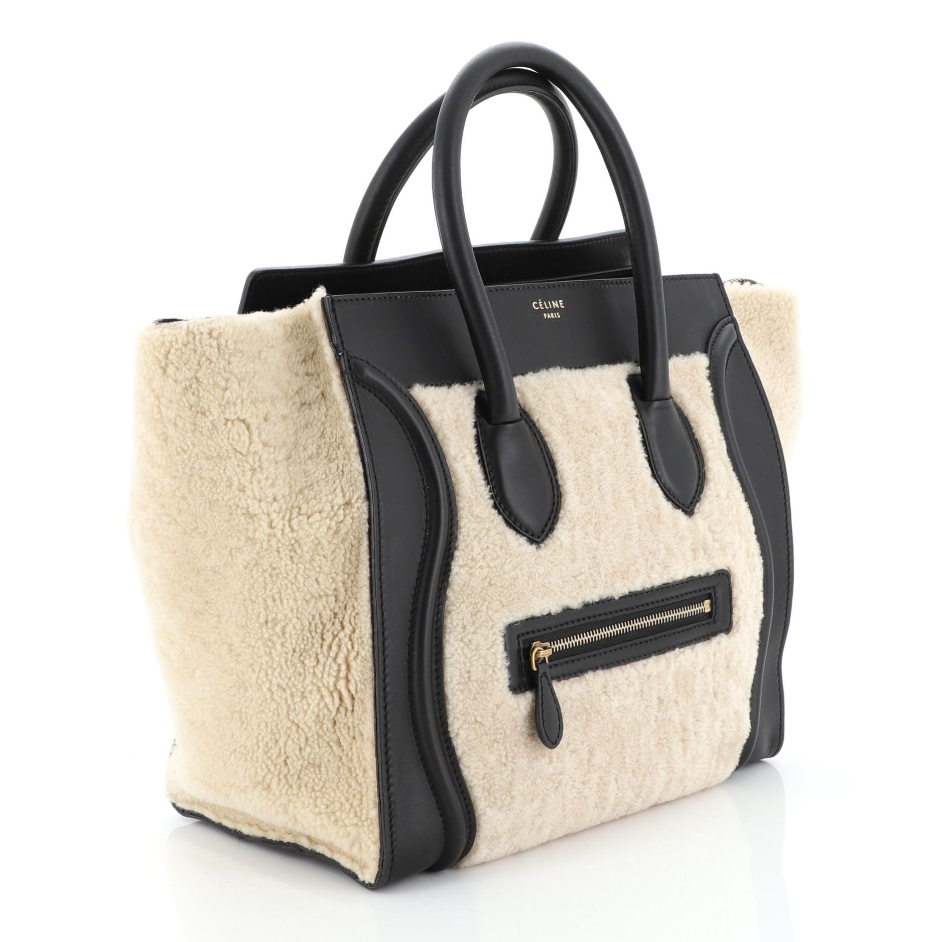 This Celine Bicolor Luggage Handbag Shearling Mini, crafted in neutral shearling and black leather, features dual rolled leather handles, front zip pocket, protective base studs, and aged gold-tone hardware. Its top zip closure opens to a black