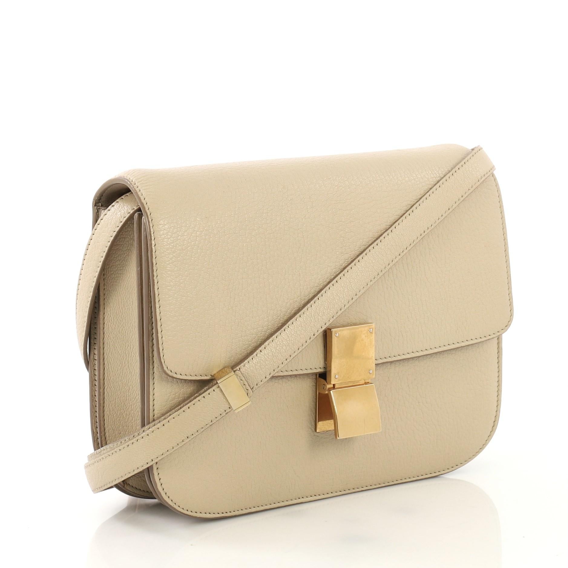 This Celine Classic Box Bag Grainy Leather Medium, crafted from beige goatskin leather, features a long adjustable strap and gold-tone hardware. Its push-tab closure opens to a beige leather interior with zip compartment and two open compartments