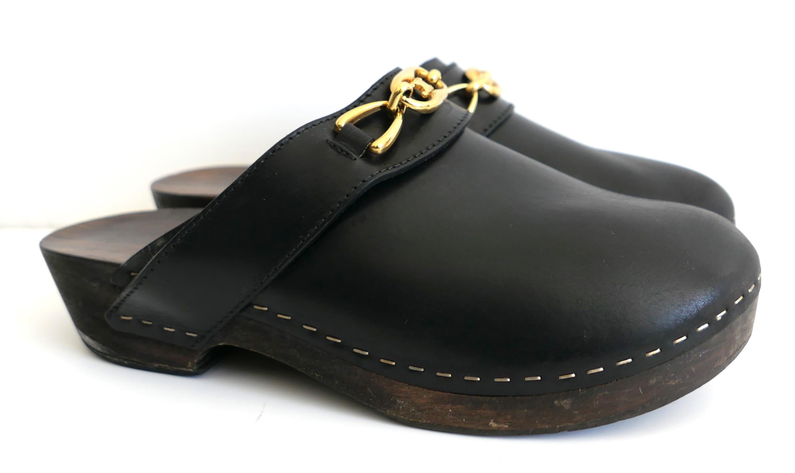 Gli iconici zoccoli Celine Les Boi. Realizzate in pelle di vitello nera liscia con leggere zeppe in legno di tiglio con finitura grezza e catena Celine Maillon Triomphe sul davanti. Hanno la punta arrotondata e la suola in gomma. Taglia 38. Misura