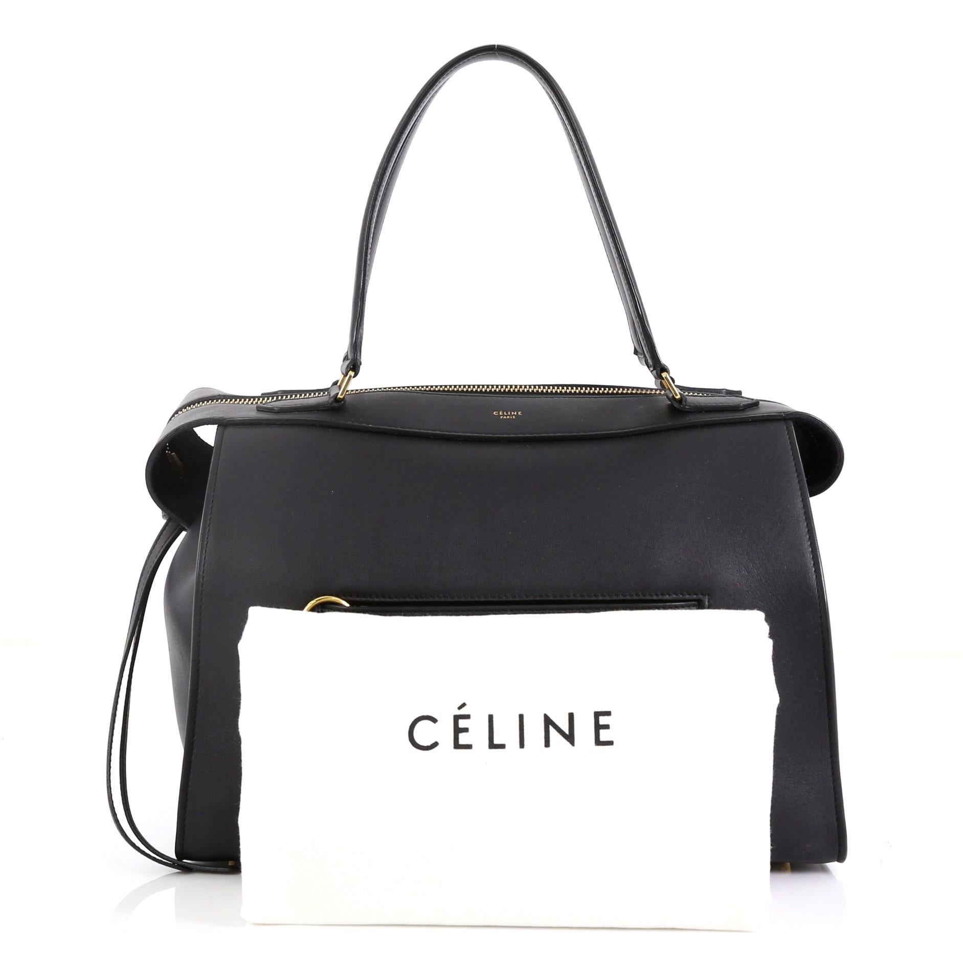 This Celine Ring Bag Leather Small, crafted in black leather, features a soft-structured silhouette, tall leather handles, ring accents on its front pocket, gold-stamped Celine logo, and gold-tone hardware. Its top zip closure opens to a black suede