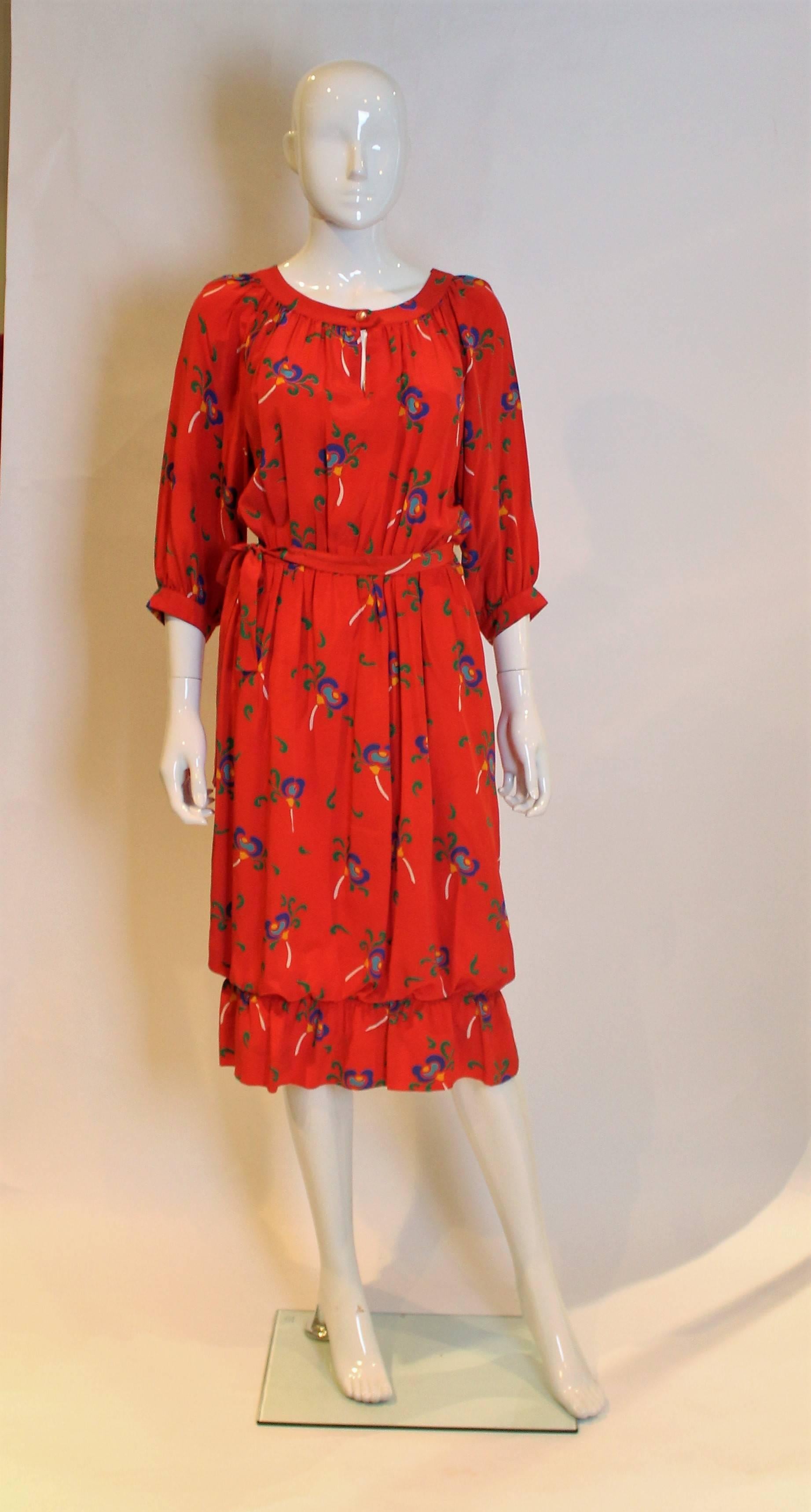 A chic silk dress by Celine.The dress has a red background with a floral print, and has a around neckline with gathering and one button opening.The dress has a side opening with poppers from the waist up and a zip opening below the waist.There is an