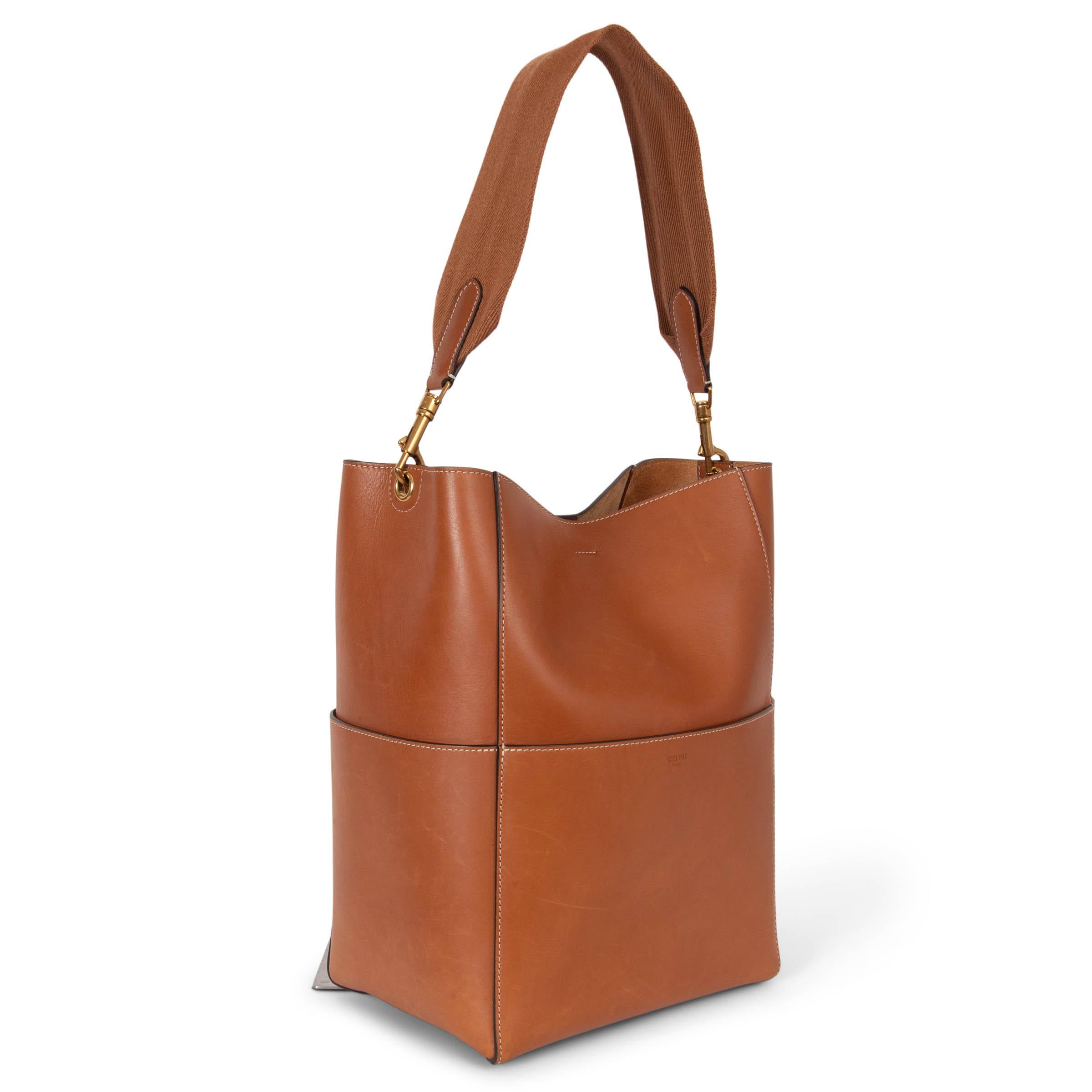 This 100% authentic Céline Sangle large bucket bag is crafted from luxurious tan smooth calfskin and accented with gold-tone hardware. It features four flat outer pockets and opens with an inner hook closure. The interior is unlined, revealing a