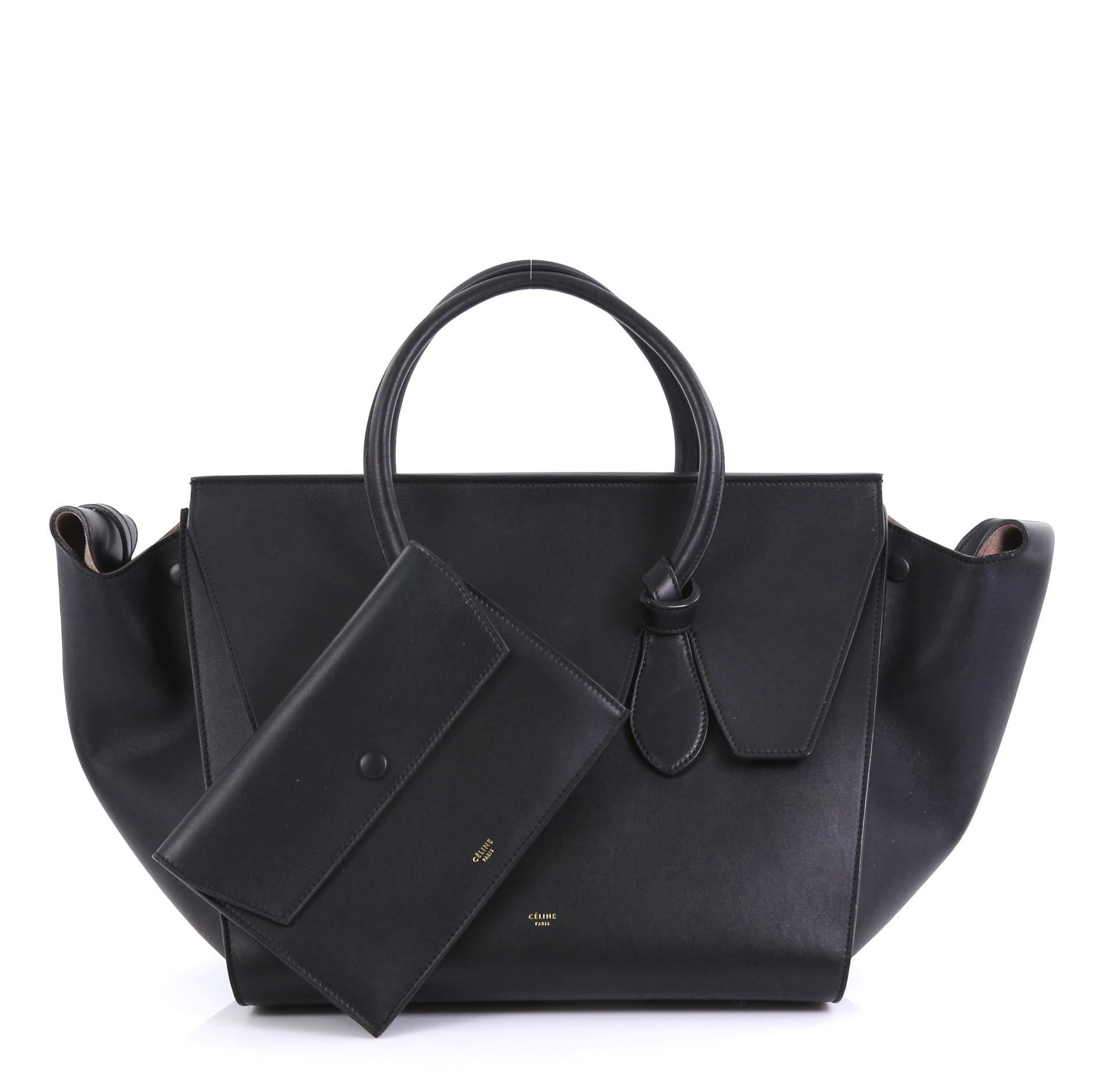 This Celine Tie Knot Tote Smooth Leather Medium, crafted from black smooth leather, features dual rolled leather handles with knot accents, protective base studs, expandable wings, and gold-tone hardware. Its flap that can be tucked inside opens to