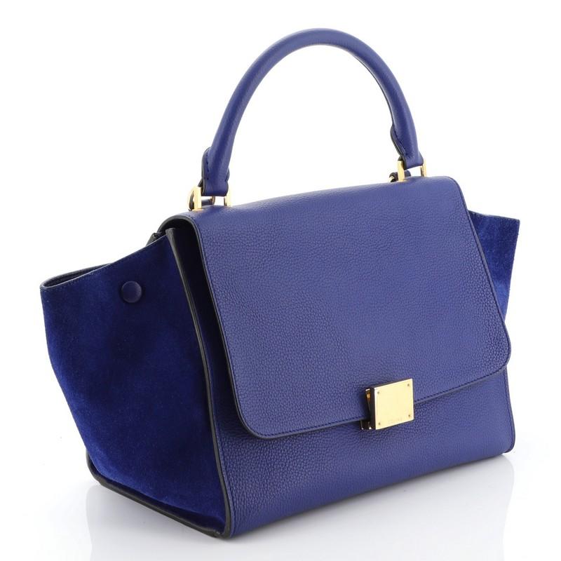 This Celine Trapeze Handbag Leather Small, crafted from black leather, features a rolled top handle, exterior back zip pocket, and gold-tone hardware. Its square flip-lock closure opens to a blue leather interior with slip pockets. 

Estimated