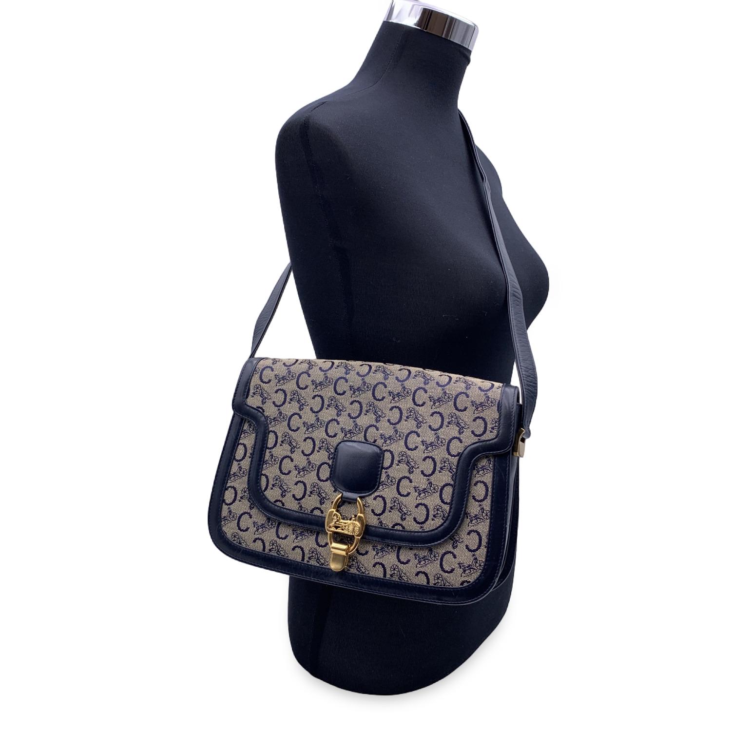 Celine 'Macadam C' box shoulder bag in Macadam C canvas with navy blue leather trim and shoulder strap. Gold metal hardware. Flap with front Caleche clasp closure. Adjustable shoulder strap. Blue leather lining. 3 side open pockets and 1 side zip