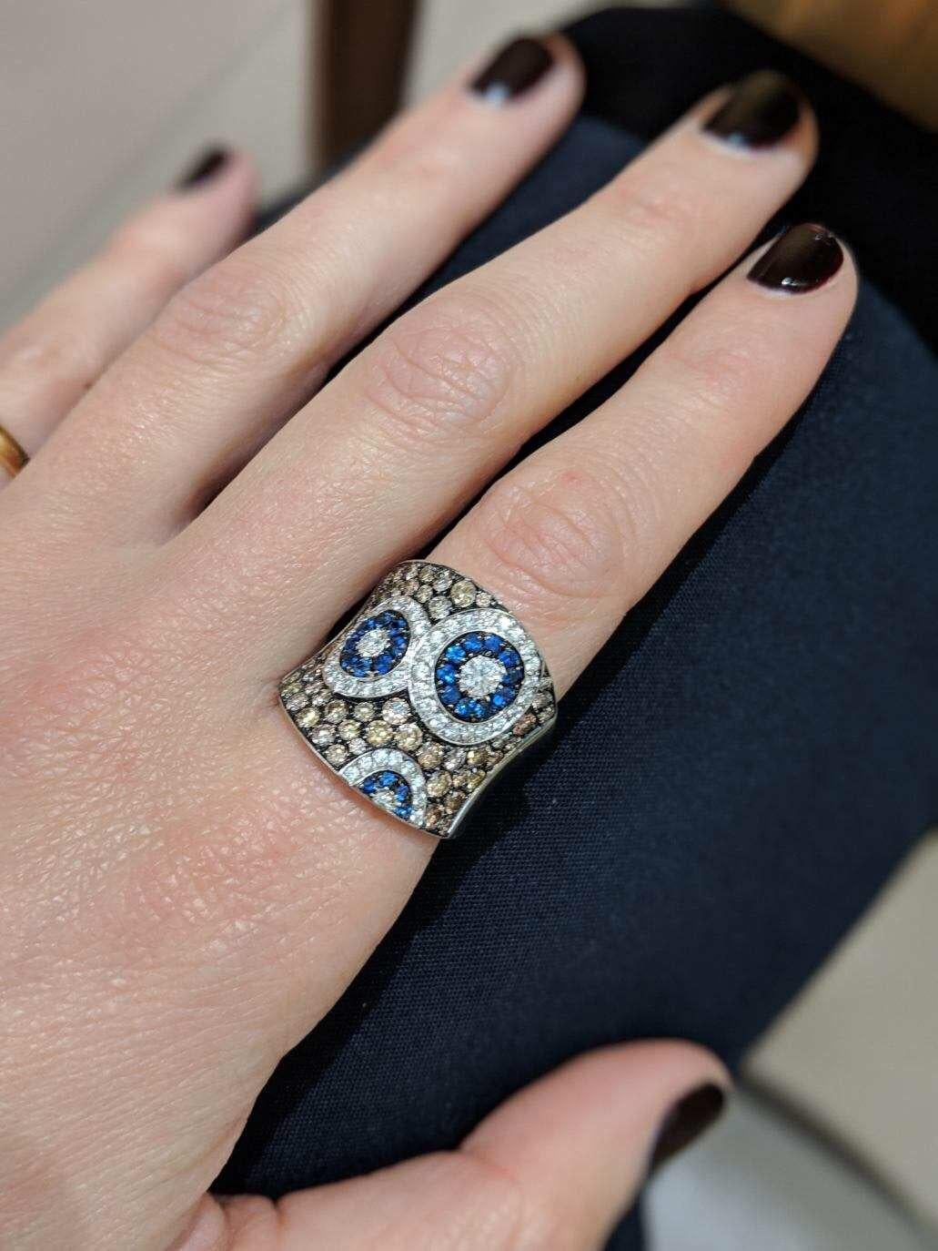 This Cellini Jewelers ring features a Kaleidoscope of white diamonds, and blue sapphire which are set within a brown diamond concave shaped ring. Set in 18 karat white gold. 
This whimsical design is part of the exclusive collection created for