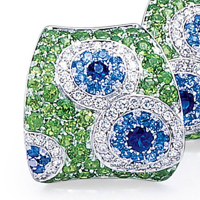 These Cellini Jewelers earrings feature a Kaleidoscope of white diamonds, and blue sapphires which are set within a green tsavorite concave shaped earring. Set in 18 karat white gold.  The earrings have a fold down post with a french clip making