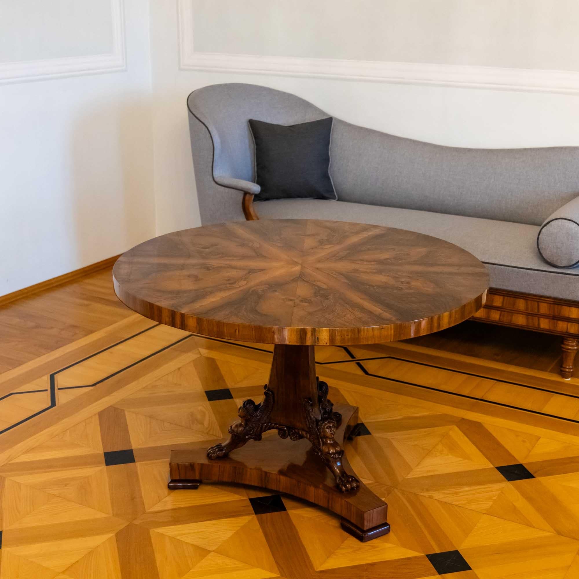 Exquisite Viennese walnut center table showcasing a round, radially veneered tabletop. The three-sided central column is adorned with carved, winged caryatids featuring lion paws. The concave base complements the central column harmoniously. This